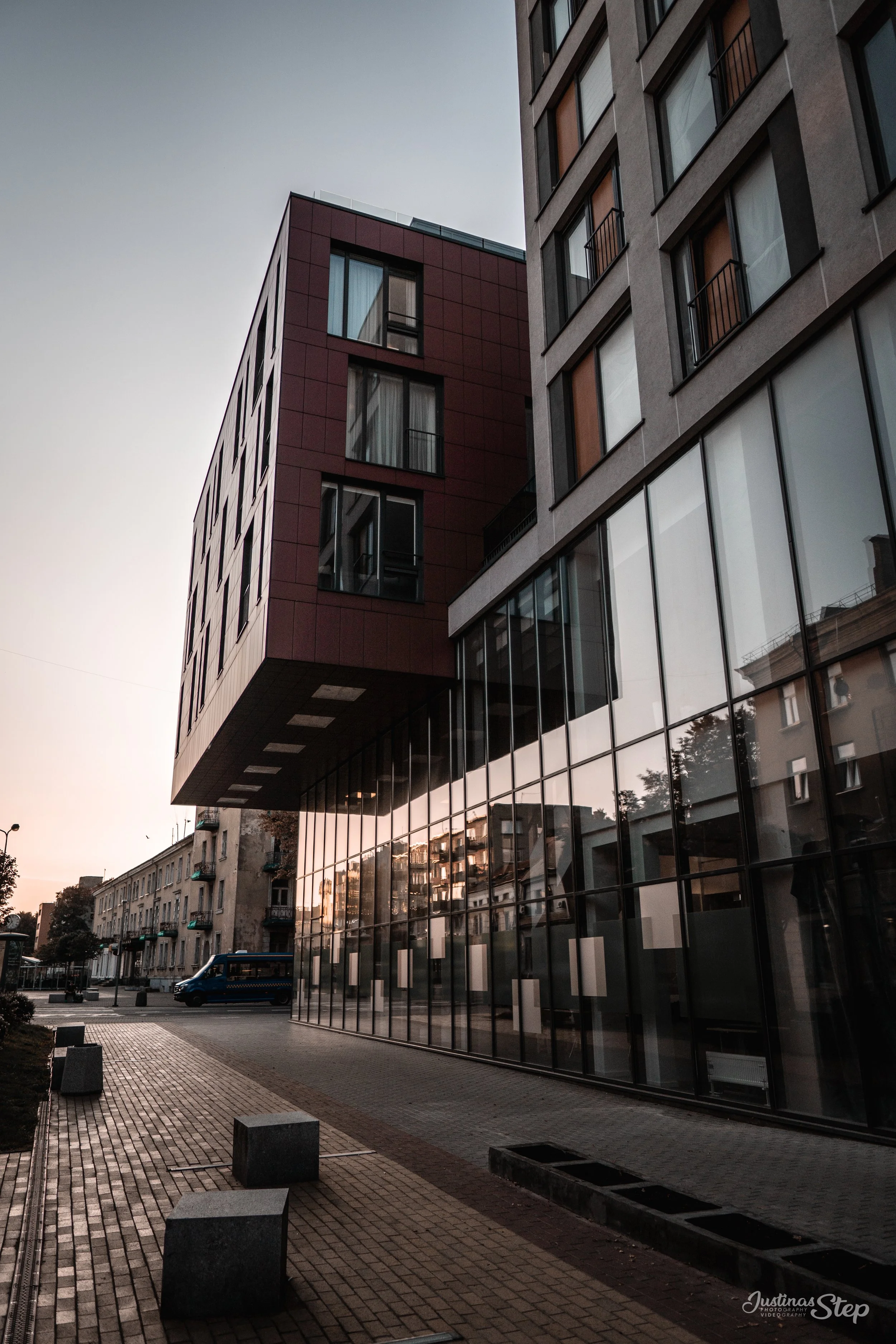 Modern multi-story building with large glass windows and a red section extending outward, reflecting surrounding buildings, with a street and benches in the foreground at sunset.