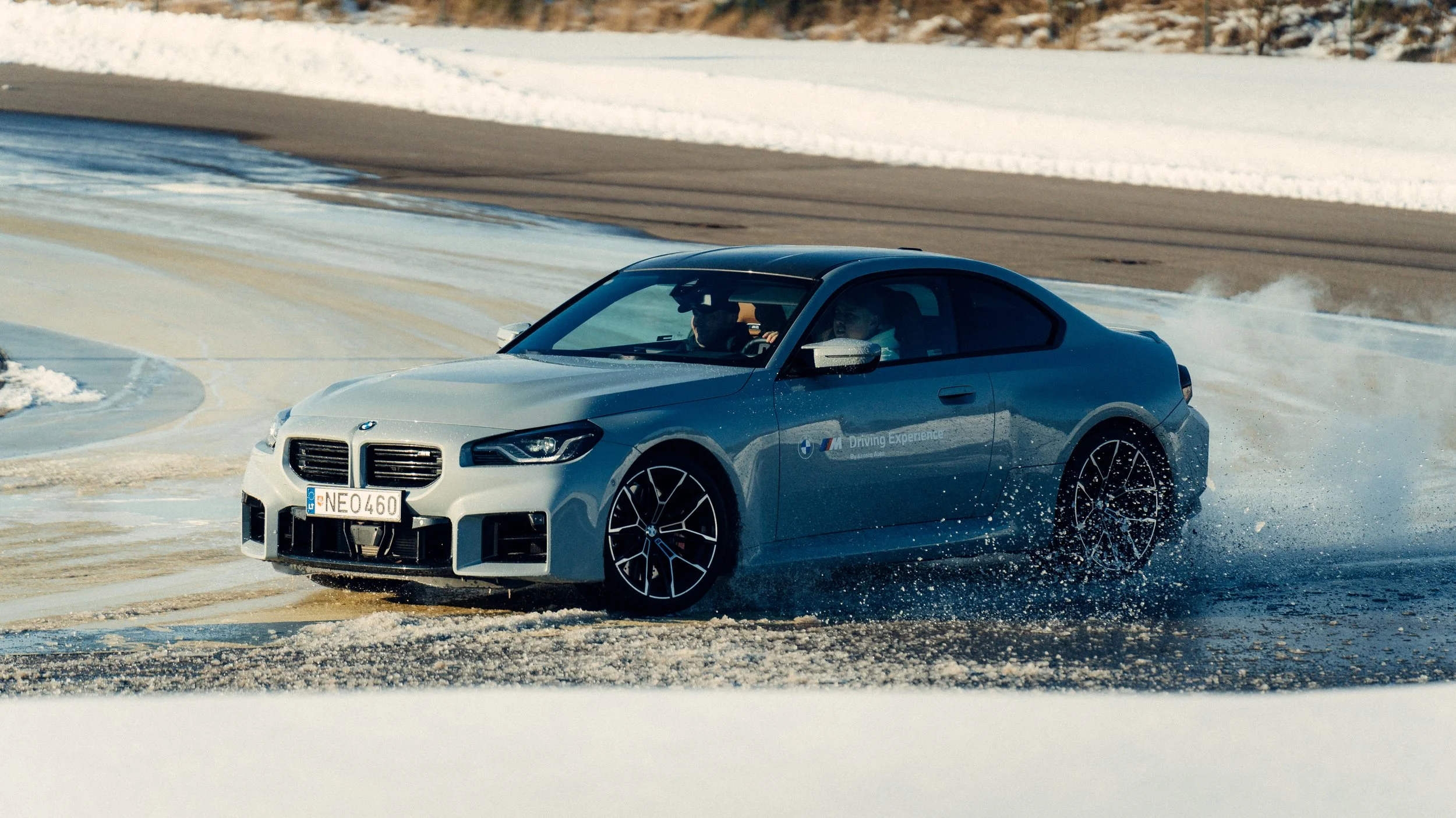 A silver BMW sports car driving on a snowy and icy track during winter, creating spray and spray of snow and ice behind it.