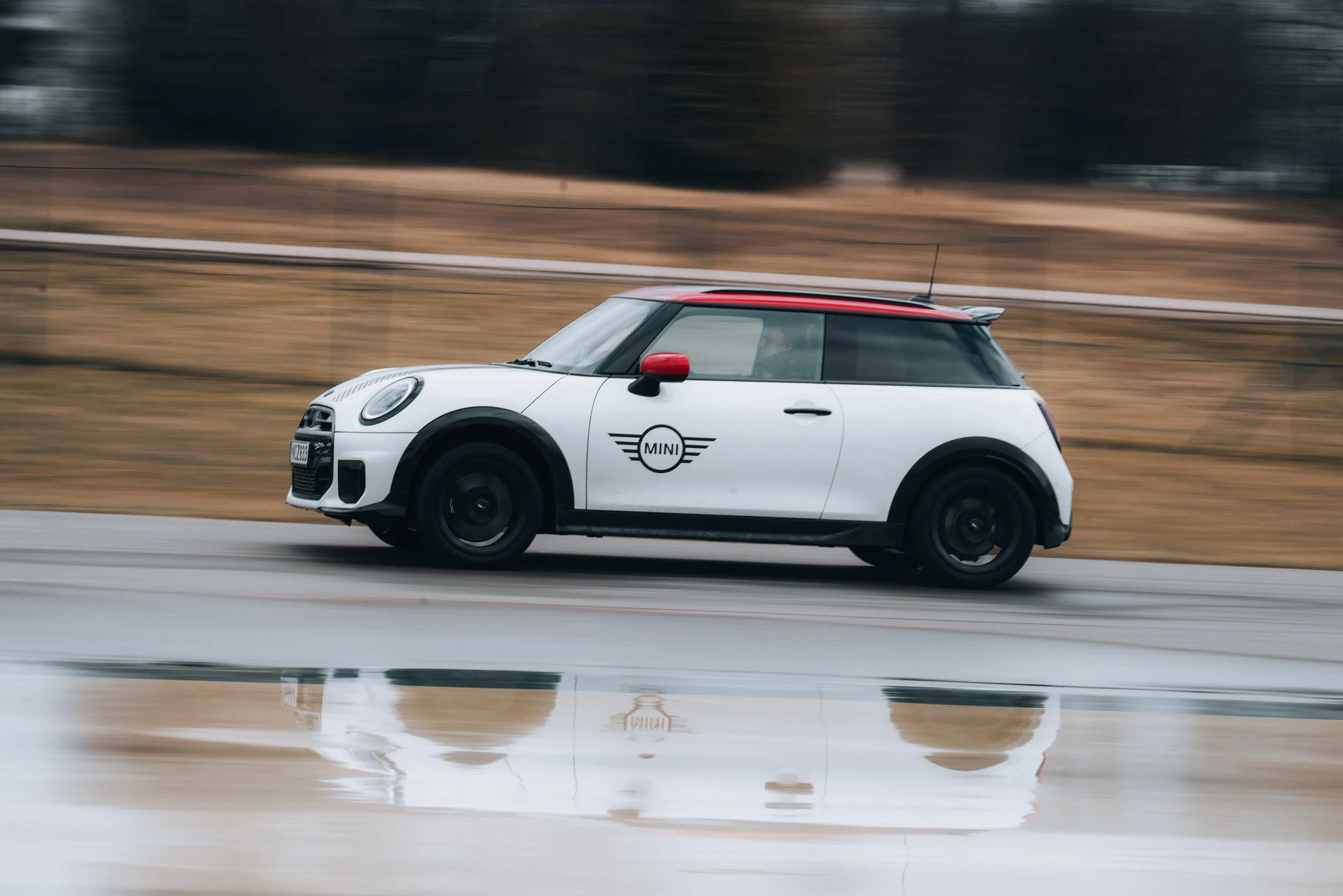 A white Mini Cooper car with black wheels and a red roof driving on a wet road, with a reflection visible on the wet surface.