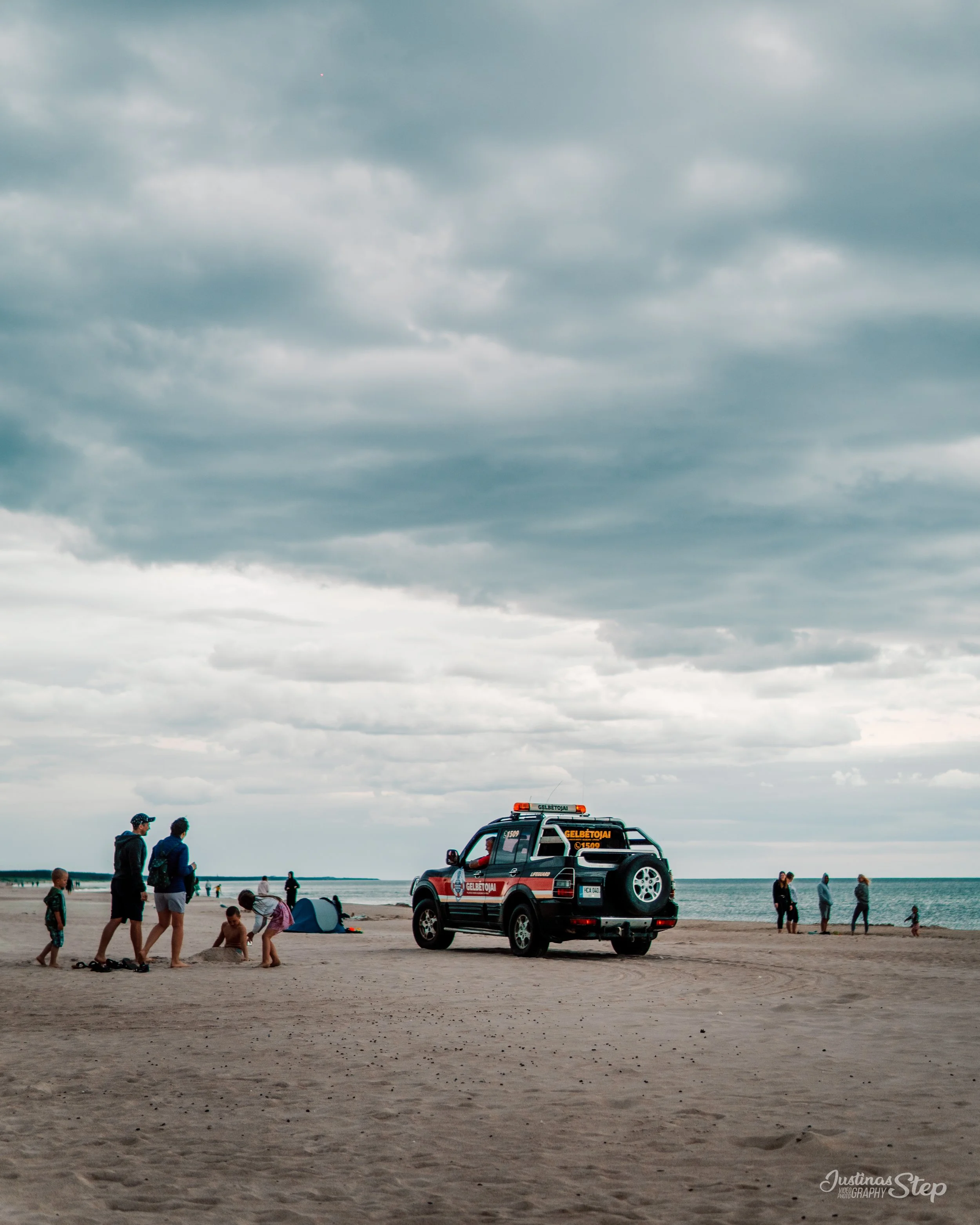 A beach scene with a police or emergency vehicle parked on the sand under a cloudy sky. Several people, including children, are playing and walking near the vehicle, with a small tent set up nearby.
