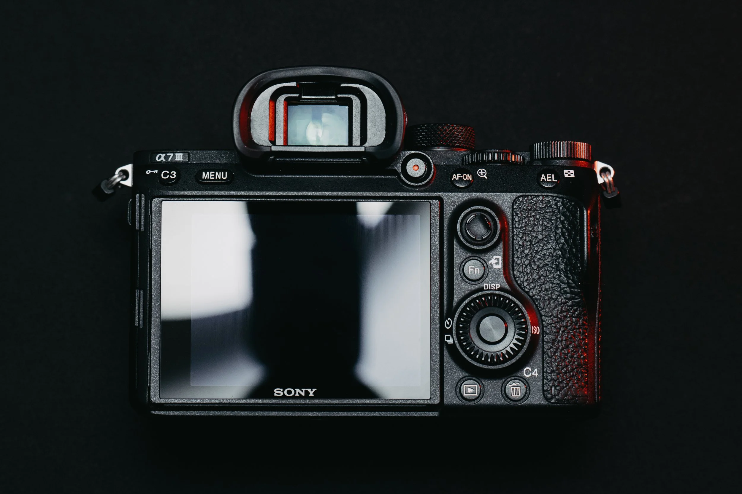 Rear view of a Sony mirrorless camera with a black textured grip, LCD screen, and various buttons and dials, against a dark background.
