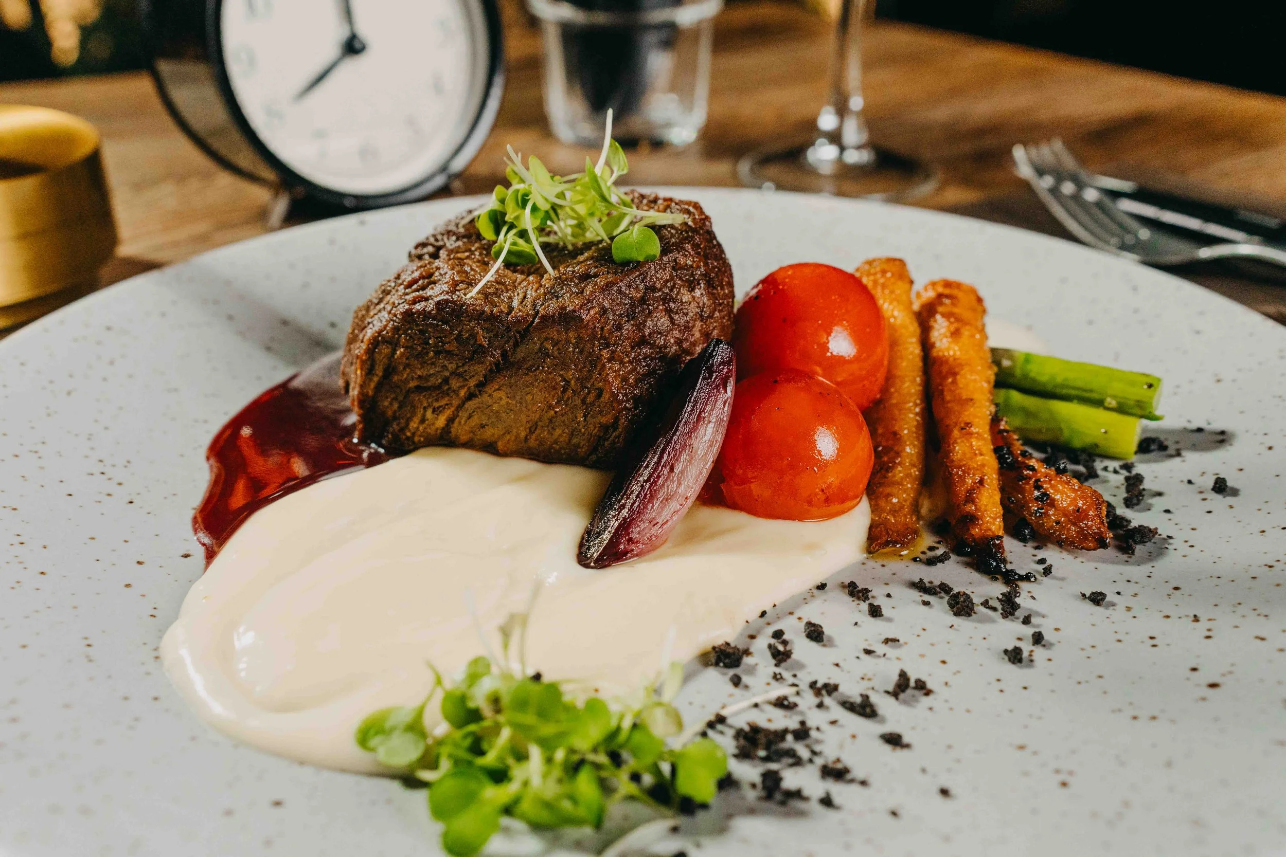A plated dish with a steak, cherry tomatoes, green beans, crispy breadsticks, and a dollop of white sauce, garnished with microgreens, with a clock and glass in the background.