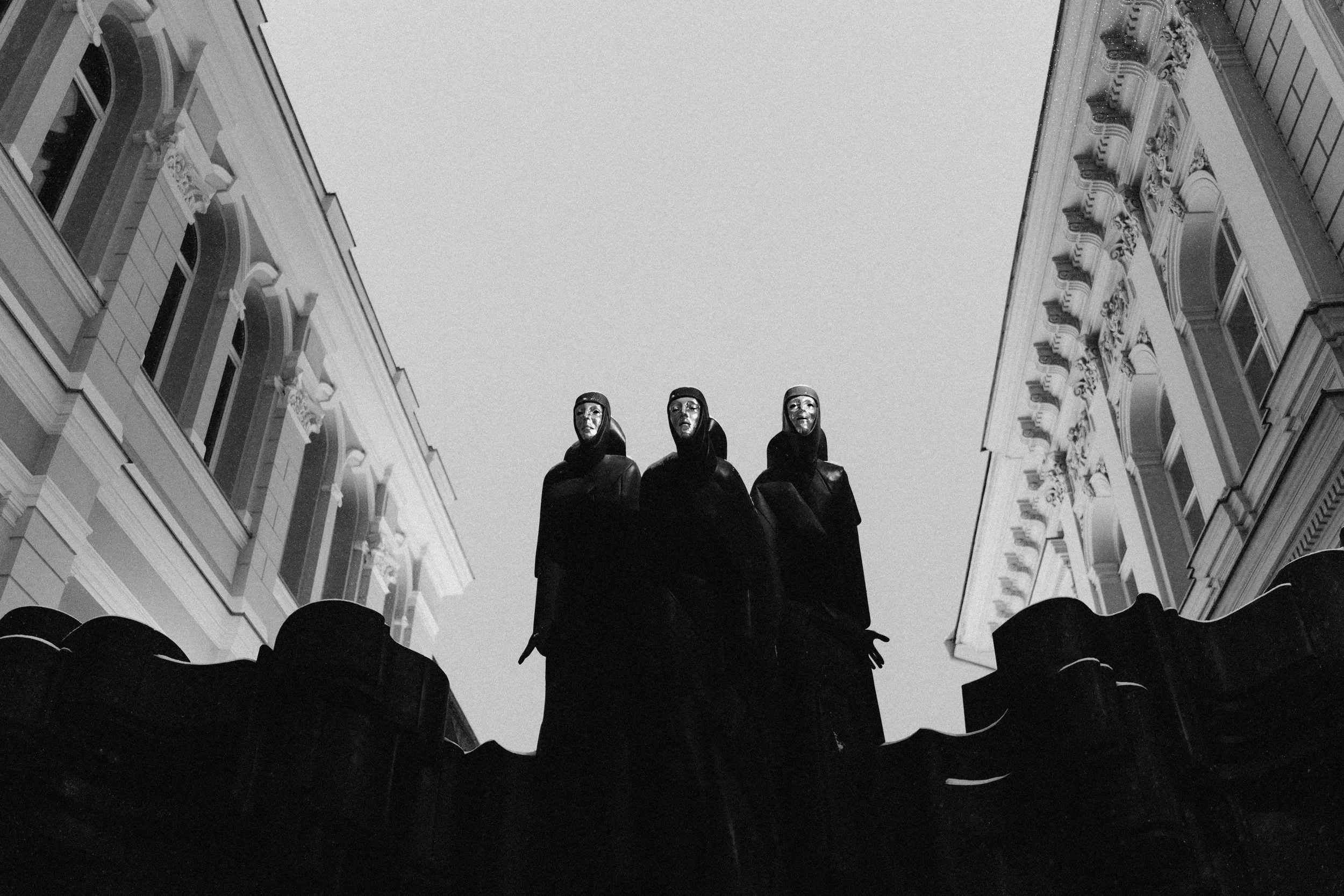 Black and white photo of three statues of robed figures with masks, positioned atop a building with ornate, classical architecture on either side.