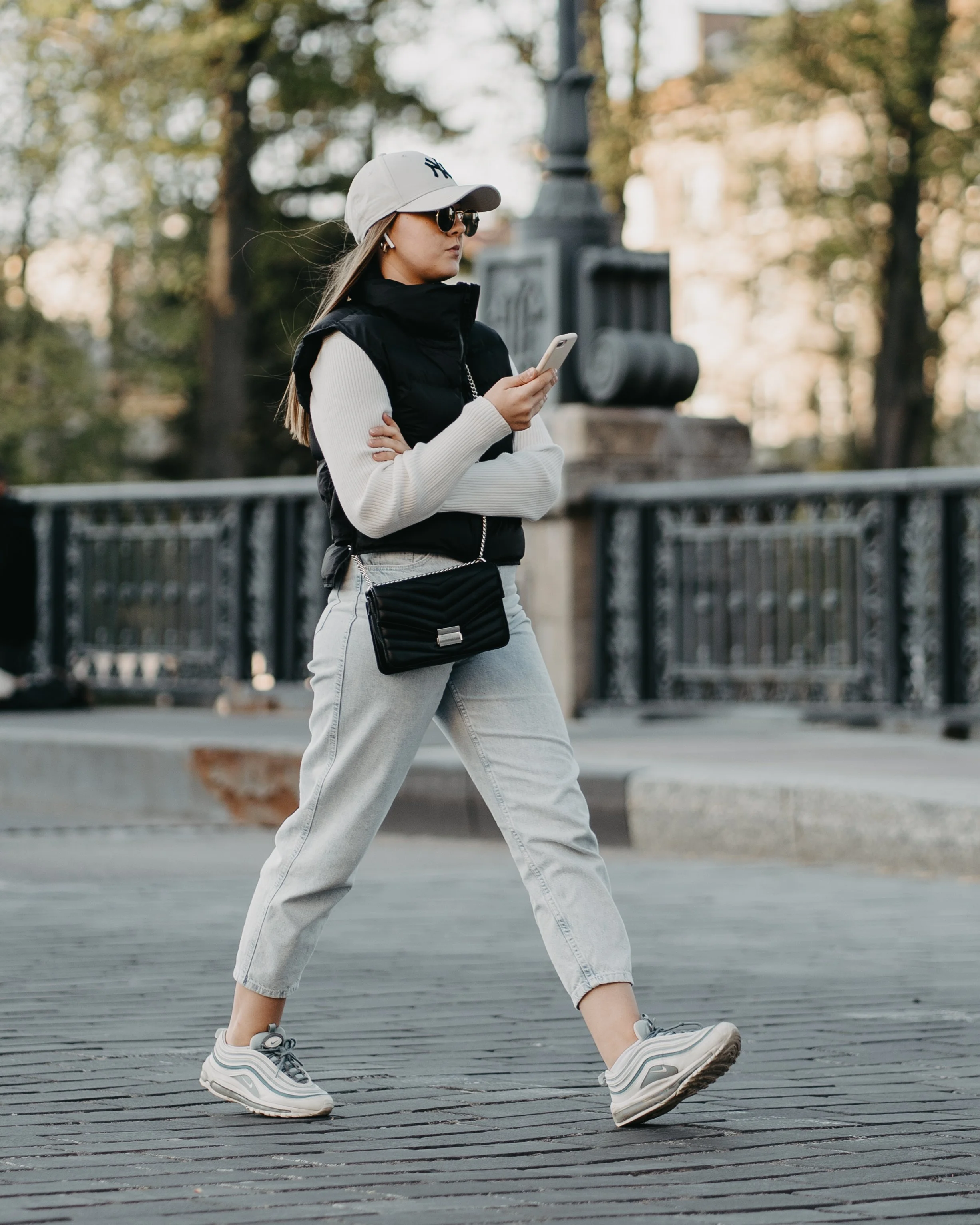 A woman walking outdoors on a city street, wearing a white cap, sunglasses, a black vest over a white sweater, light gray jeans, and sneakers, while looking at her phone.