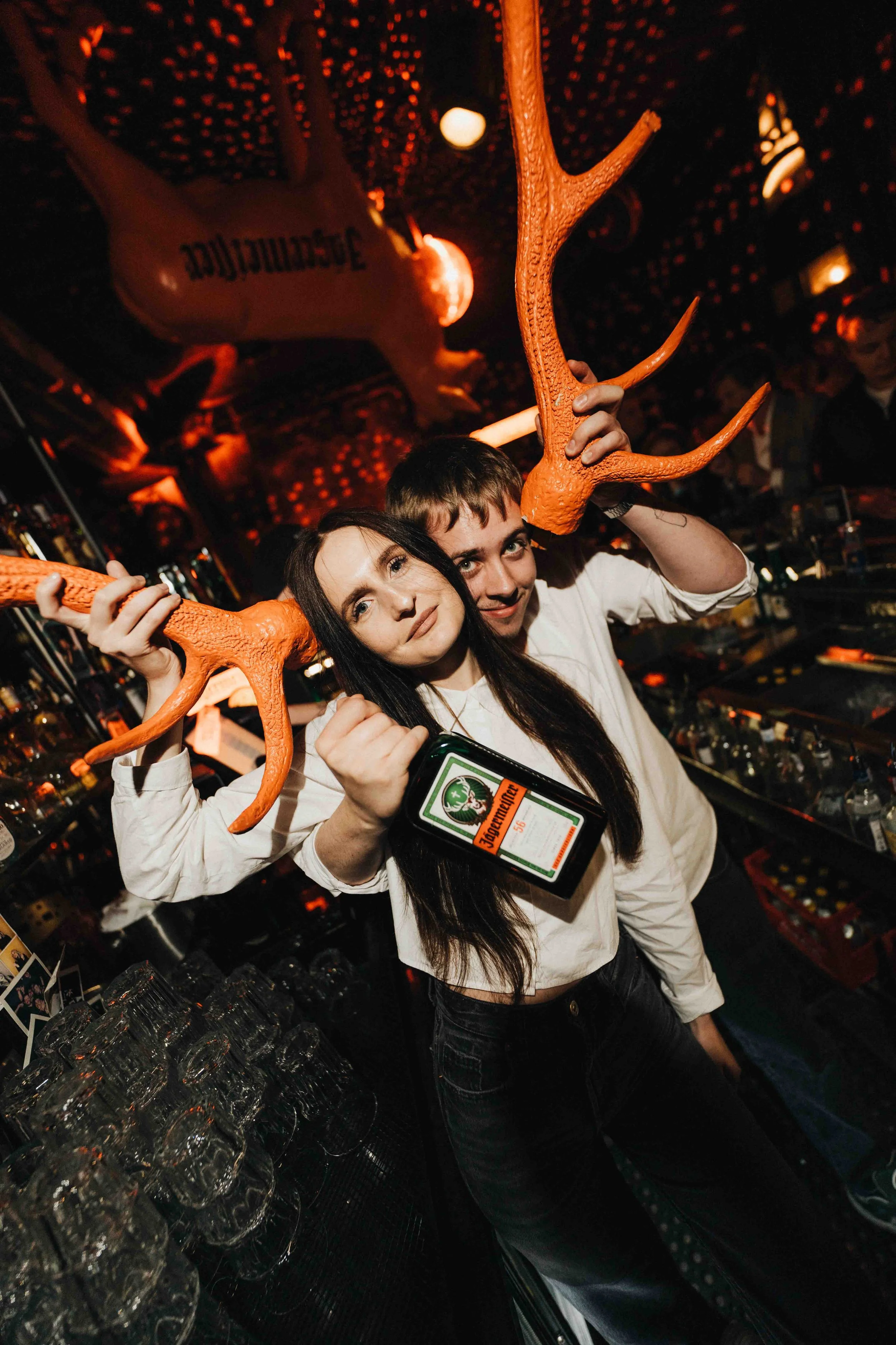 Two people, a man and a woman, are at a bar, holding large orange antler-shaped decorations, with the woman also holding a smartphone displaying a Jägermeister advertisement. The bar is dimly lit with a festive atmosphere, and there are shelves stock
