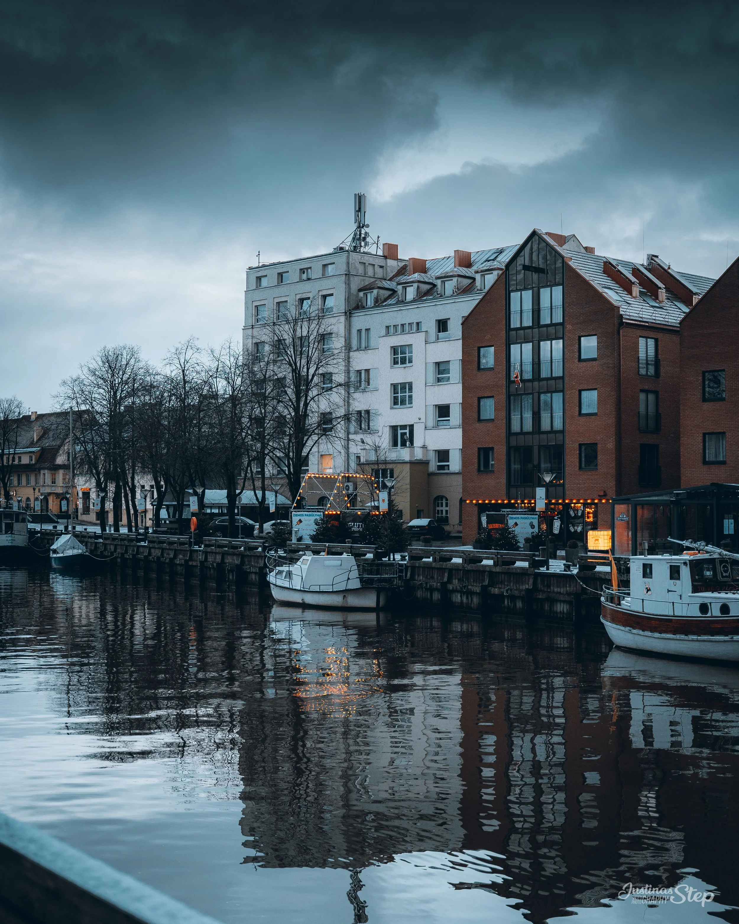 Photograph of a harbor with boats docked along the water, and multi-story buildings behind, under dark stormy clouds.