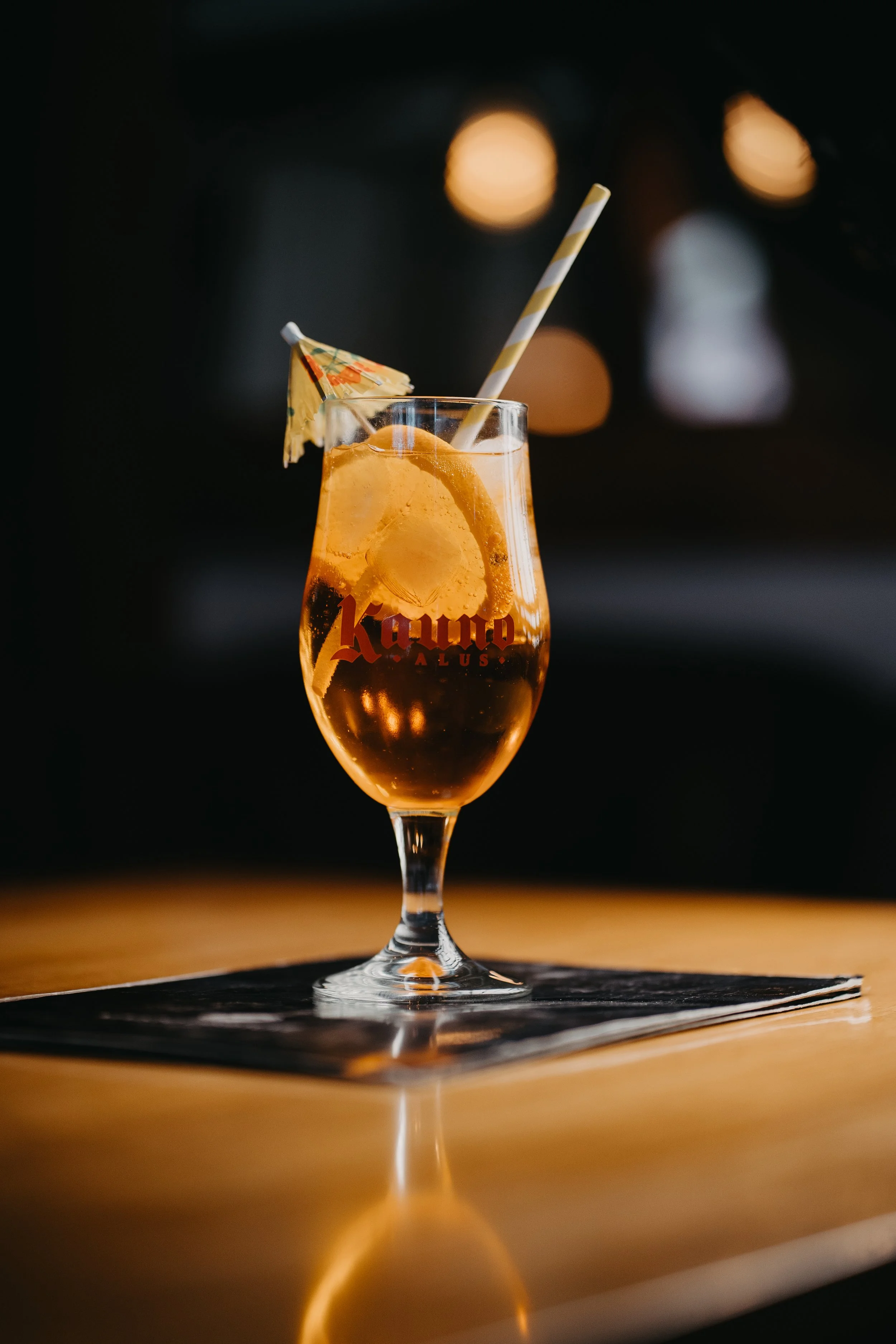 A tall glass of beer with a wedge of lemon, a cocktail umbrella, and a striped straw, placed on a wooden surface at an indoor bar or restaurant.