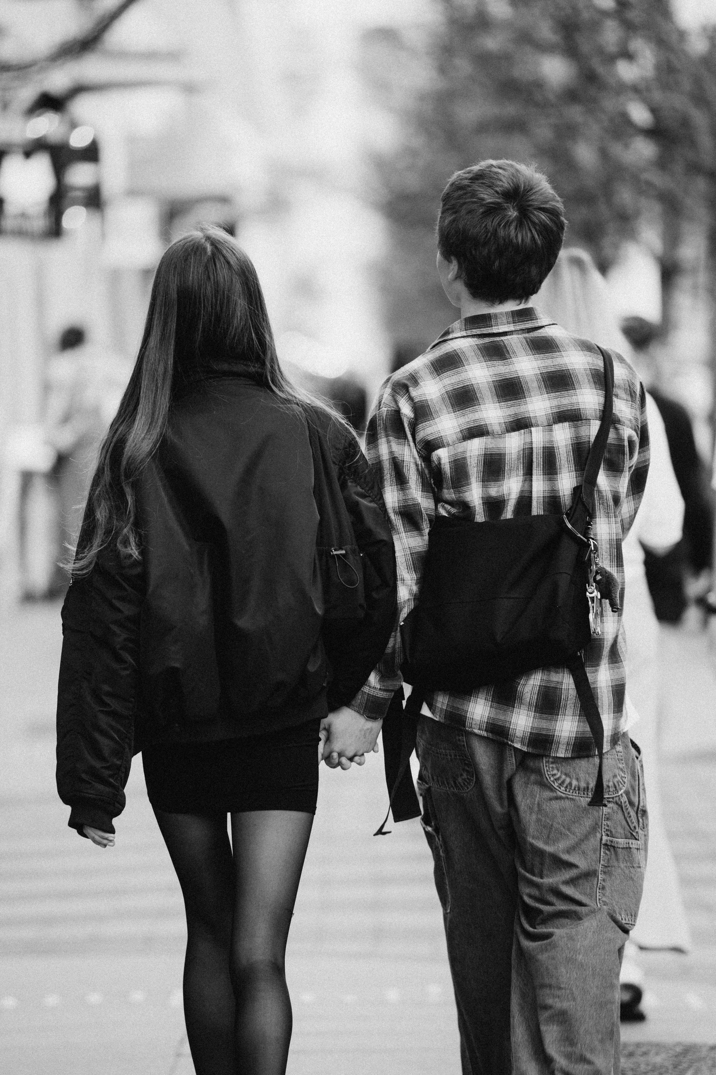 A young couple holding hands and walking down a city street, seen from behind. The woman has long hair and is wearing a jacket with a skirt and tights, while the man has short hair, a plaid shirt, and is carrying a shoulder bag.
