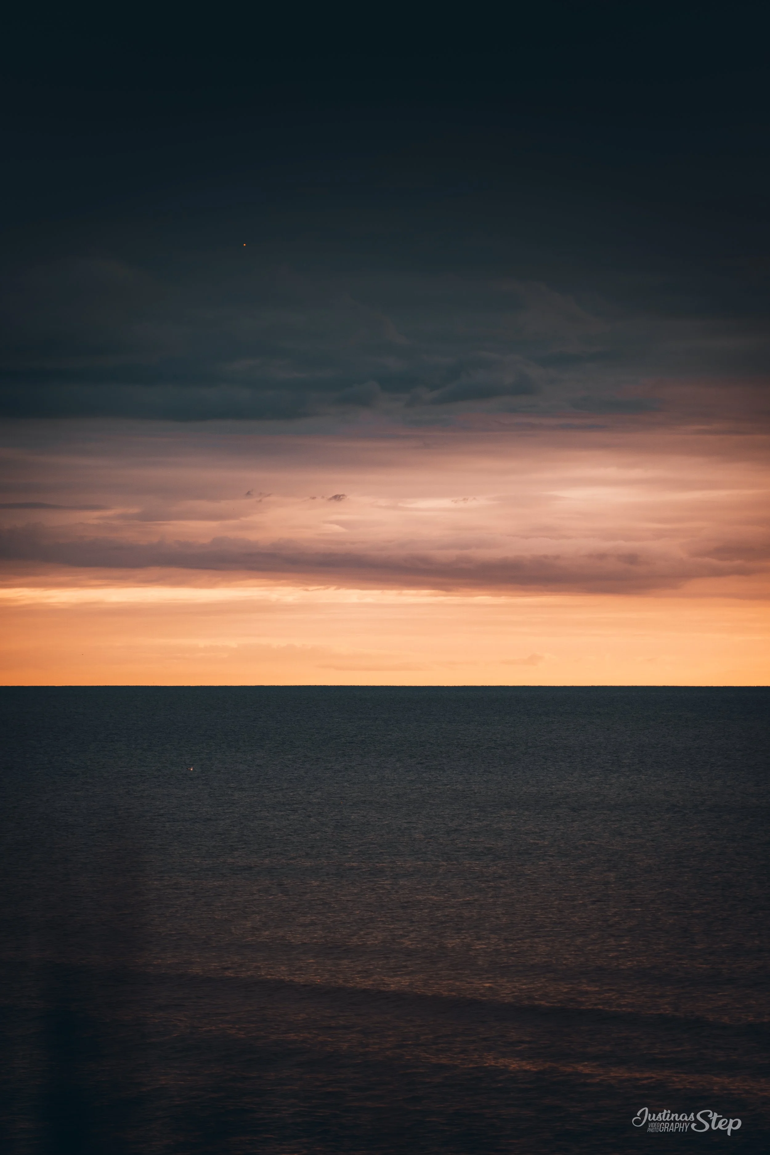 A sunset over the ocean with a partly cloudy sky, featuring dark clouds and a pinkish-orange horizon.