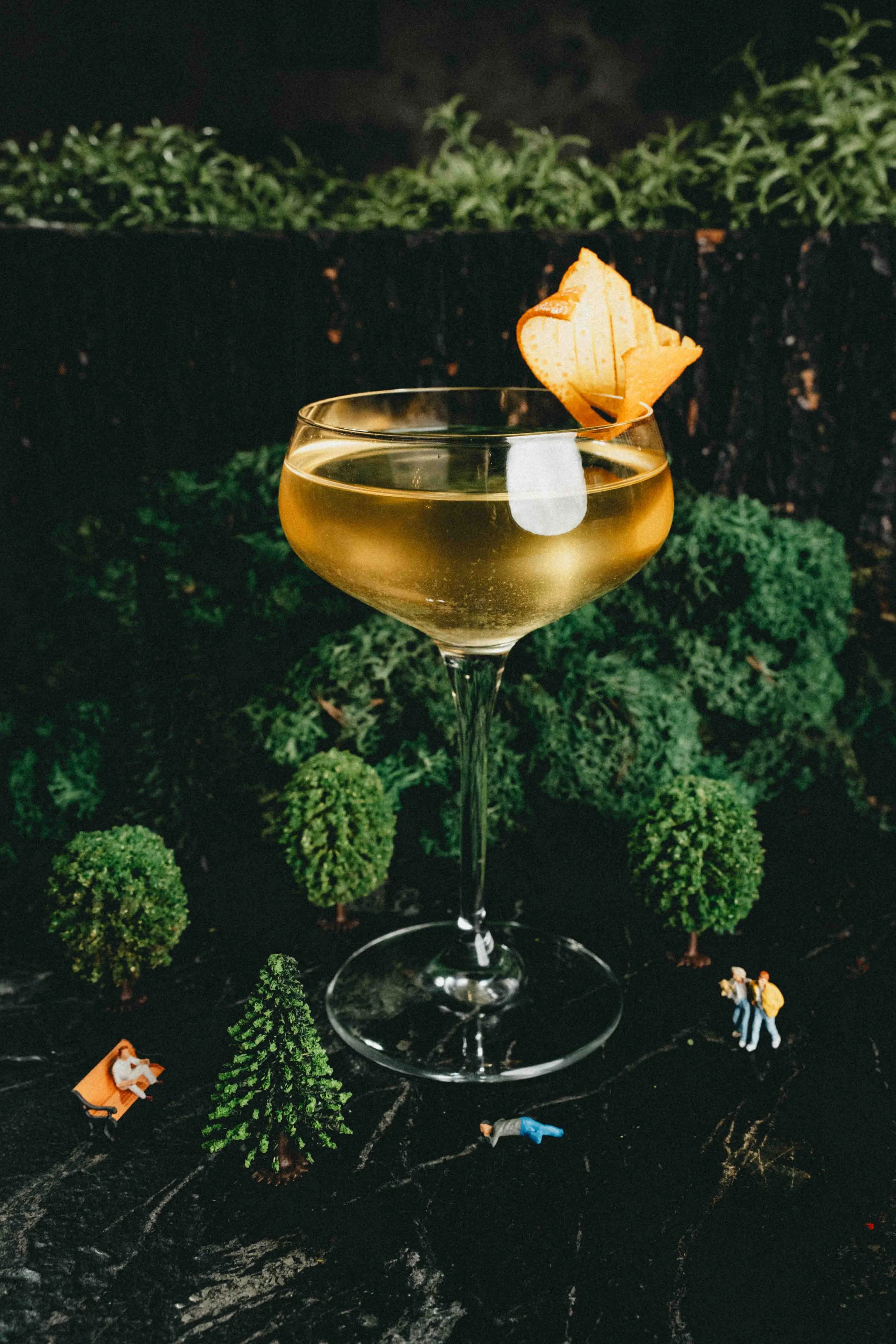 A cocktail in a coupe glass with a lemon peel garnish, set against a backdrop of miniature trees and tiny figurines of people in a miniature park scene.