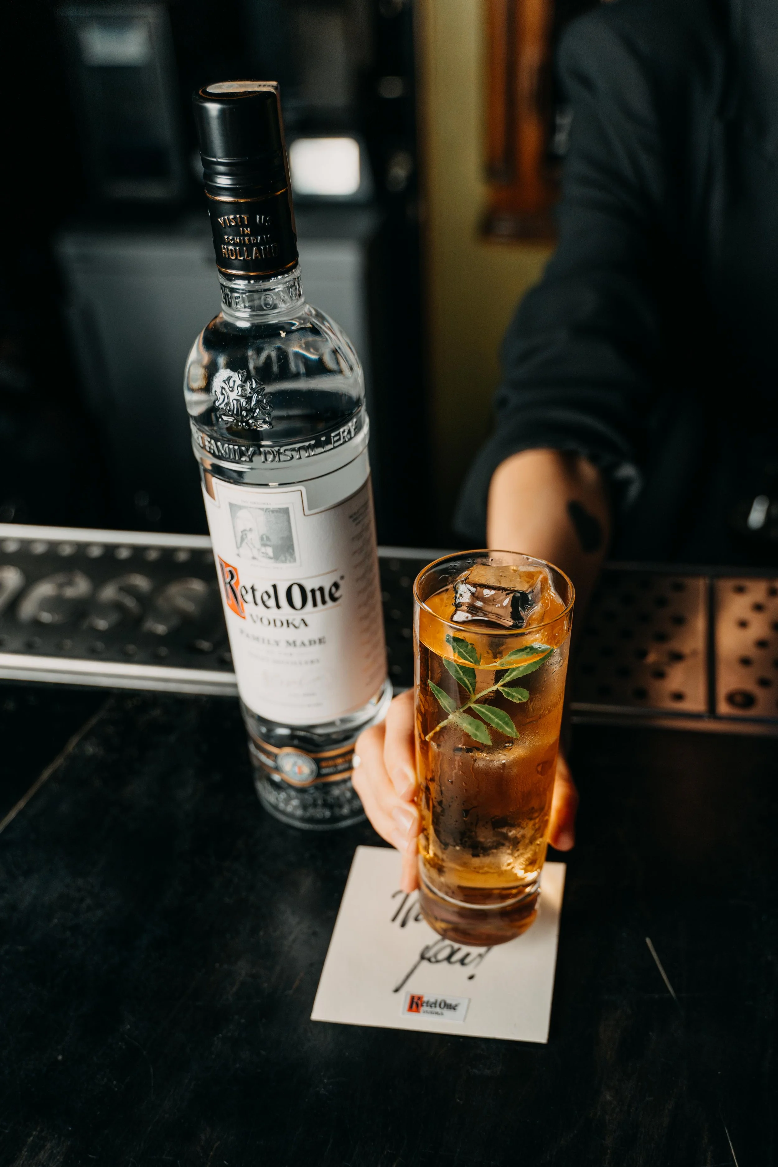 A bartender holding a glass of iced cocktail with mint leaves and ice cubes, next to a bottle of Ketel One vodka on a bar counter.