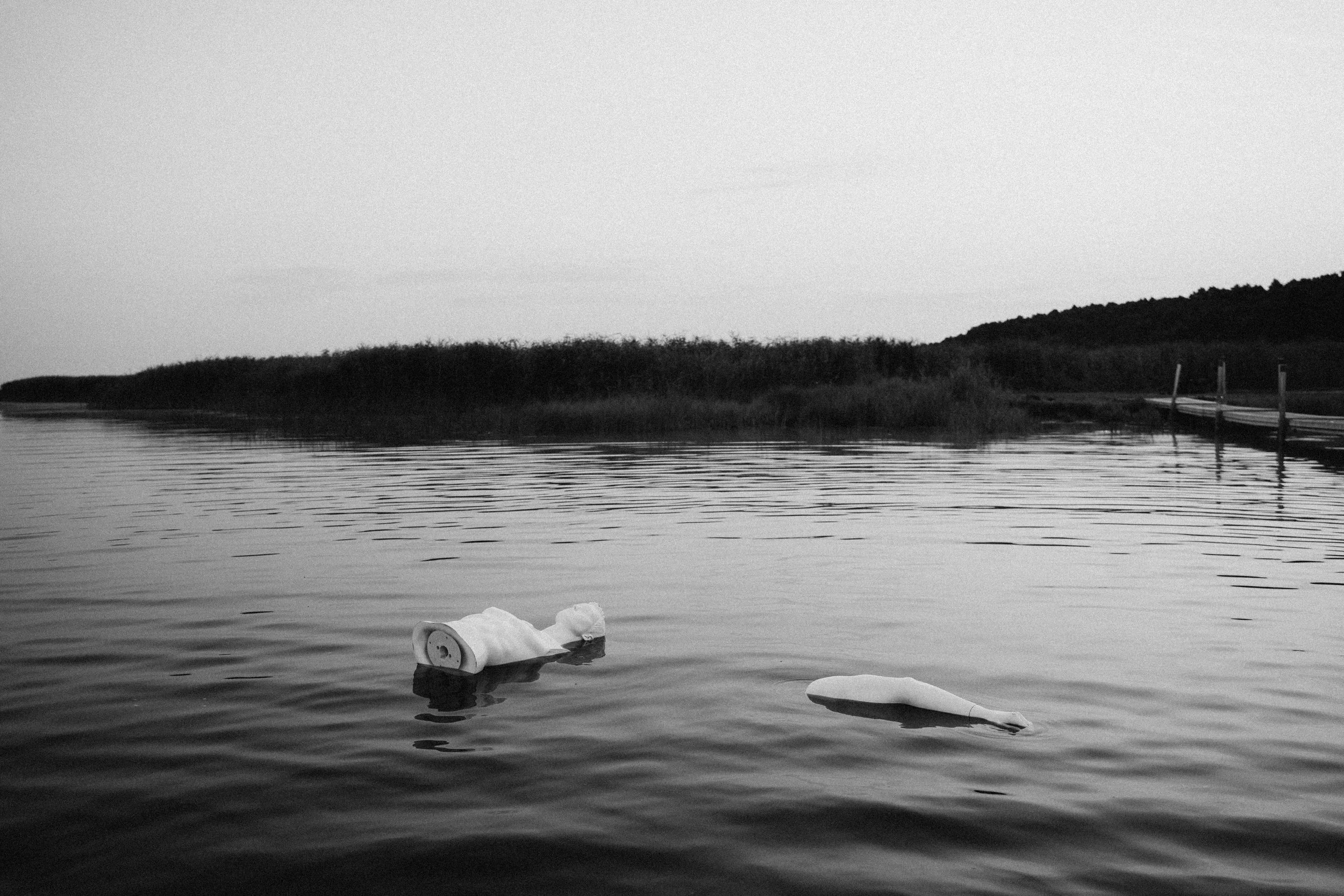 Black and white photo of a body of water with two partially submerged objects, one resembling a rolled-up blanket or fabric and the other an elongated object, near the shoreline with trees in the background and a dock on the right side.