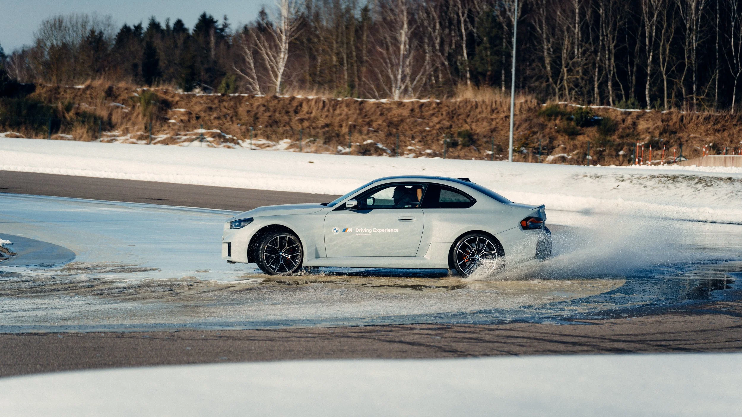 A silver BMW sports car driving on a snowy test track, creating a spray of snow and water, with a wooded hillside in the background.