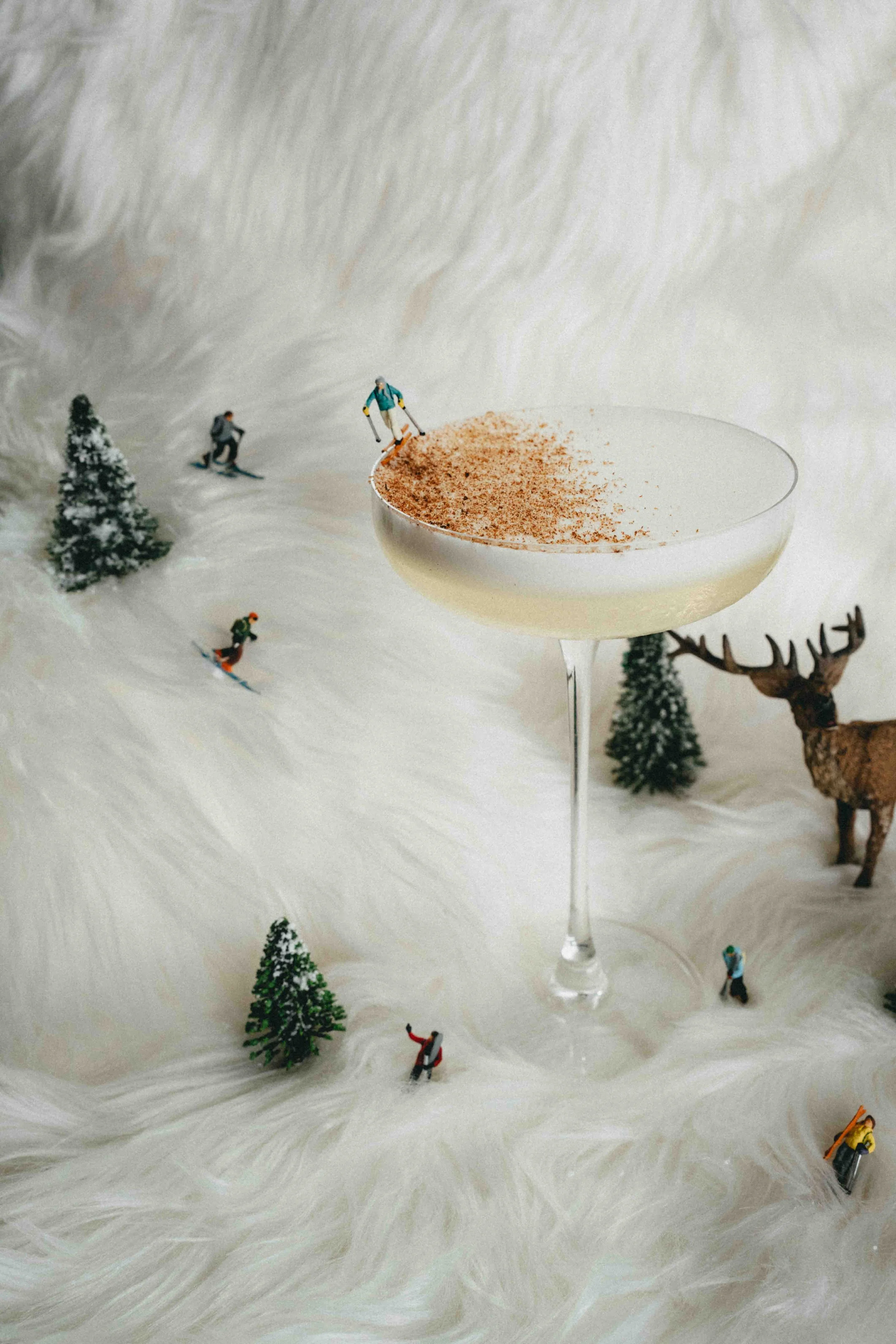 Miniature winter scene with skiers and a reindeer, surrounding a large cocktail glass with a creamy drink topped with cinnamon powder.