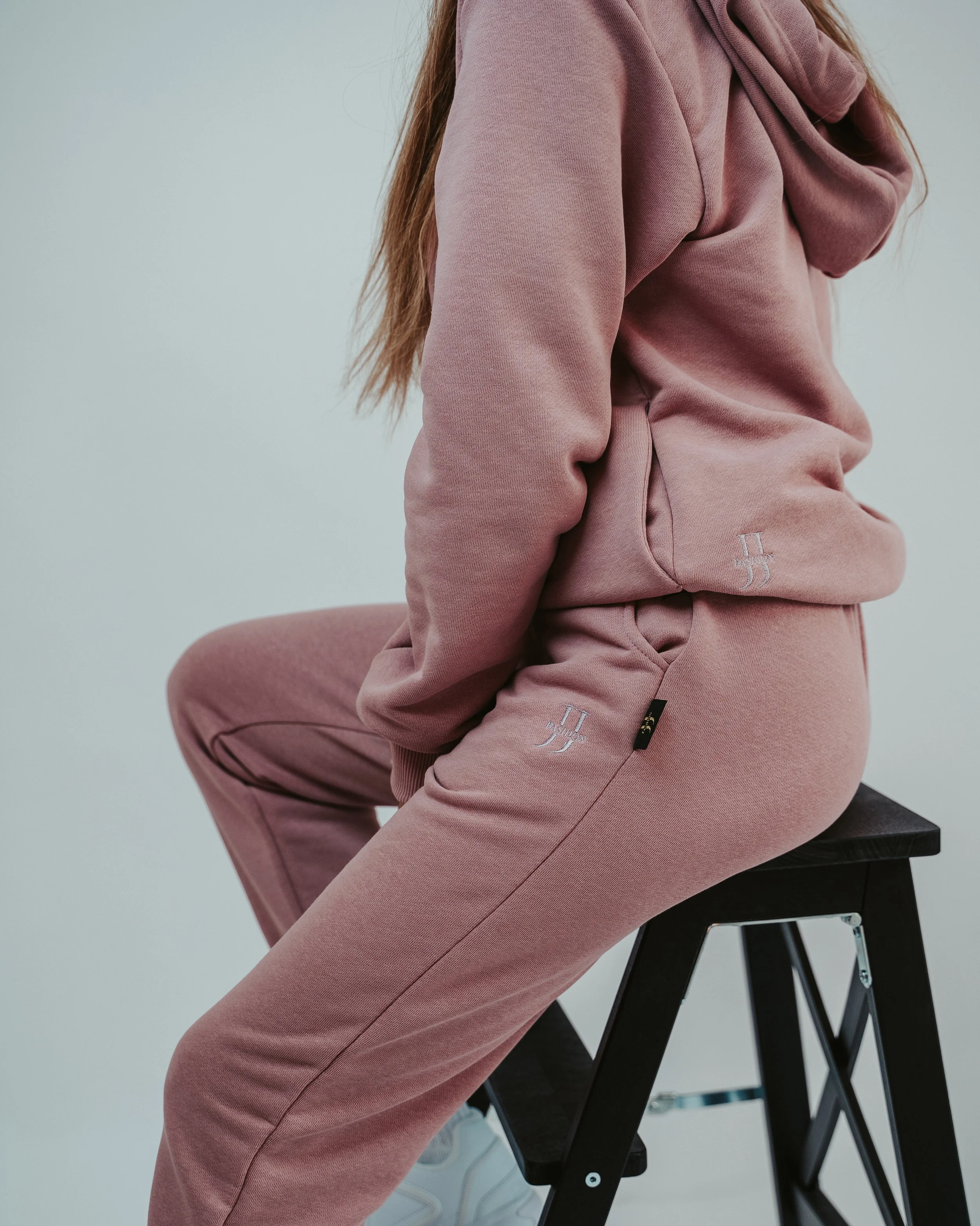 Person wearing a pink hoodie and matching sweatpants sitting on a black stool against a plain light background.
