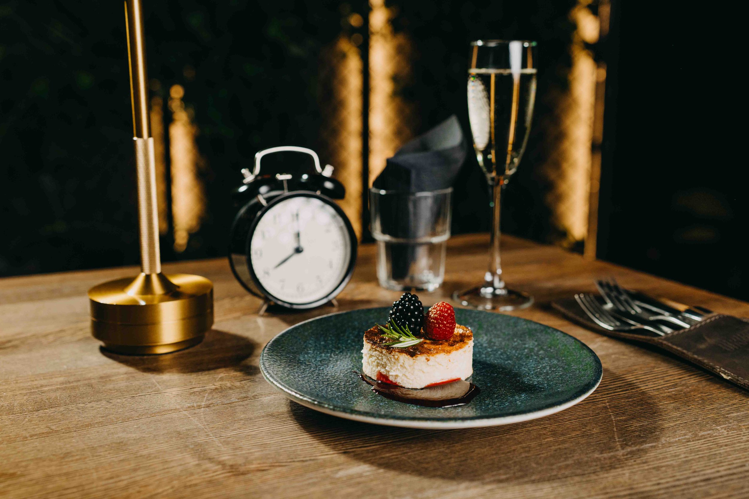 A slice of cheesecake with blackberries and raspberries on top, drizzled with chocolate sauce, on a dark green plate on a wooden table, with a glass of champagne, a clock, and a candle holder in the background.