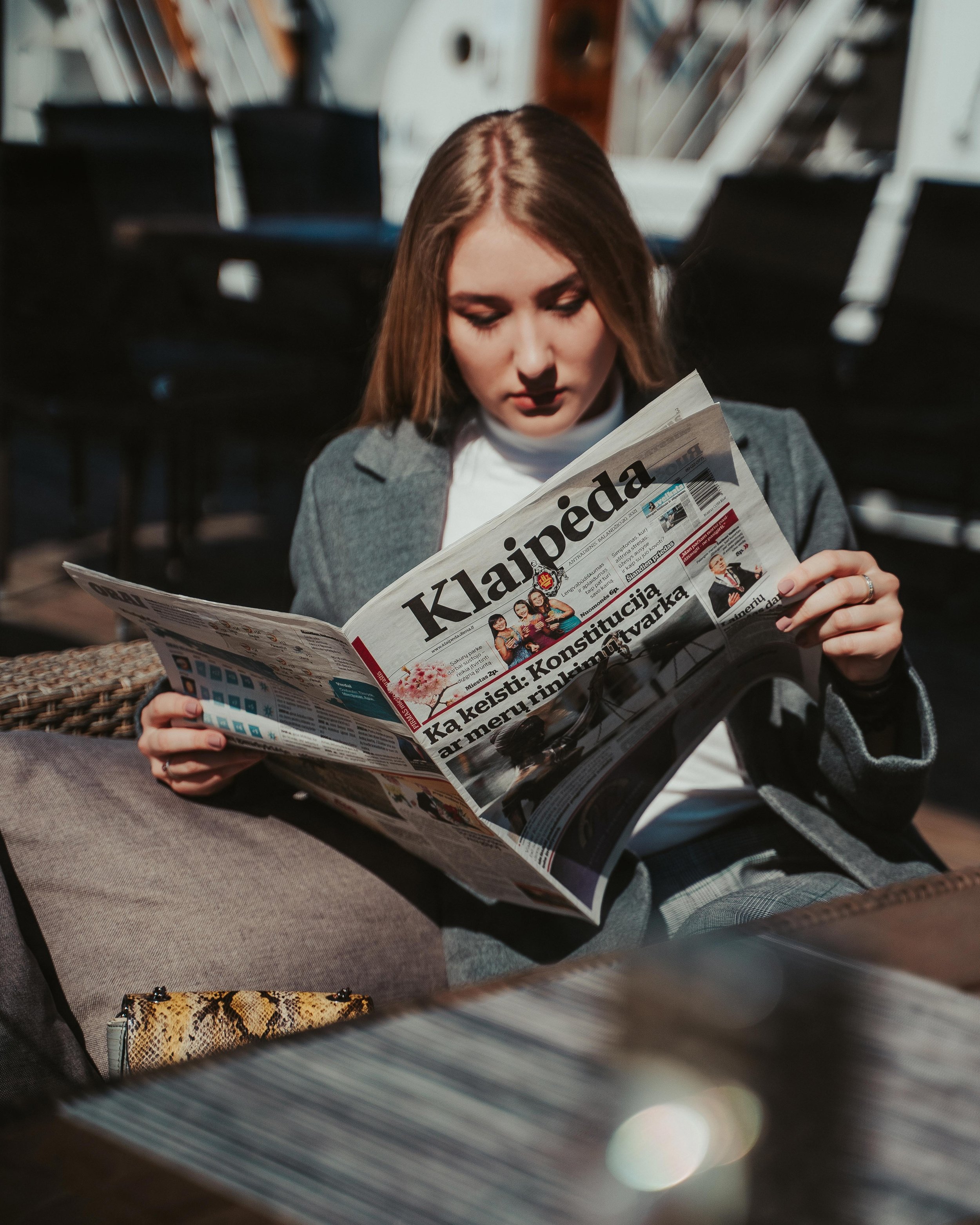 A young woman with long brown hair reading a newspaper outdoors on a sunny day, sitting at a table with a patterned bag and a glass in the foreground.