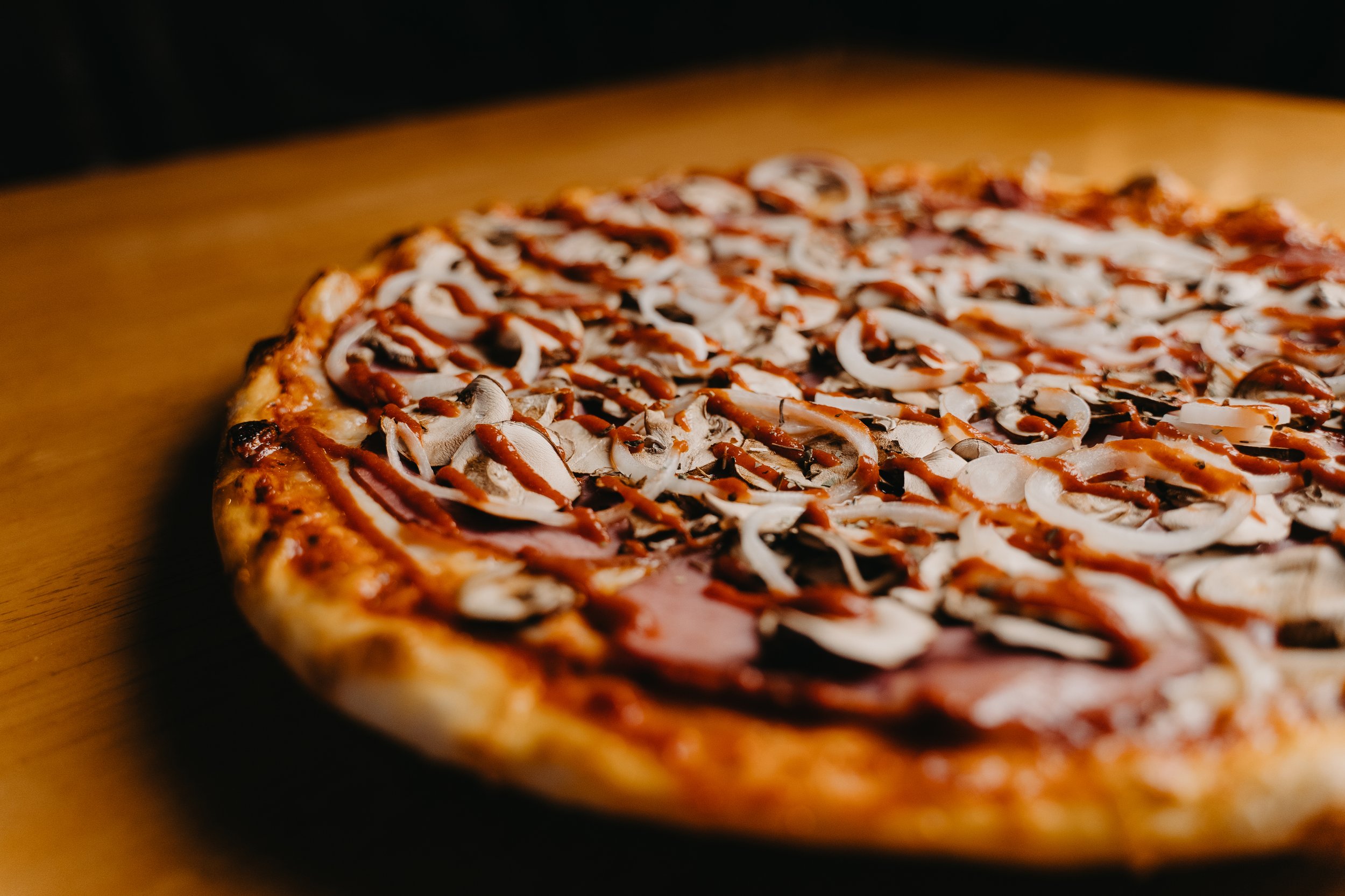 Close-up of a freshly baked pizza with mushrooms, onions, pepperoni, and drizzled sauce on a wooden surface.
