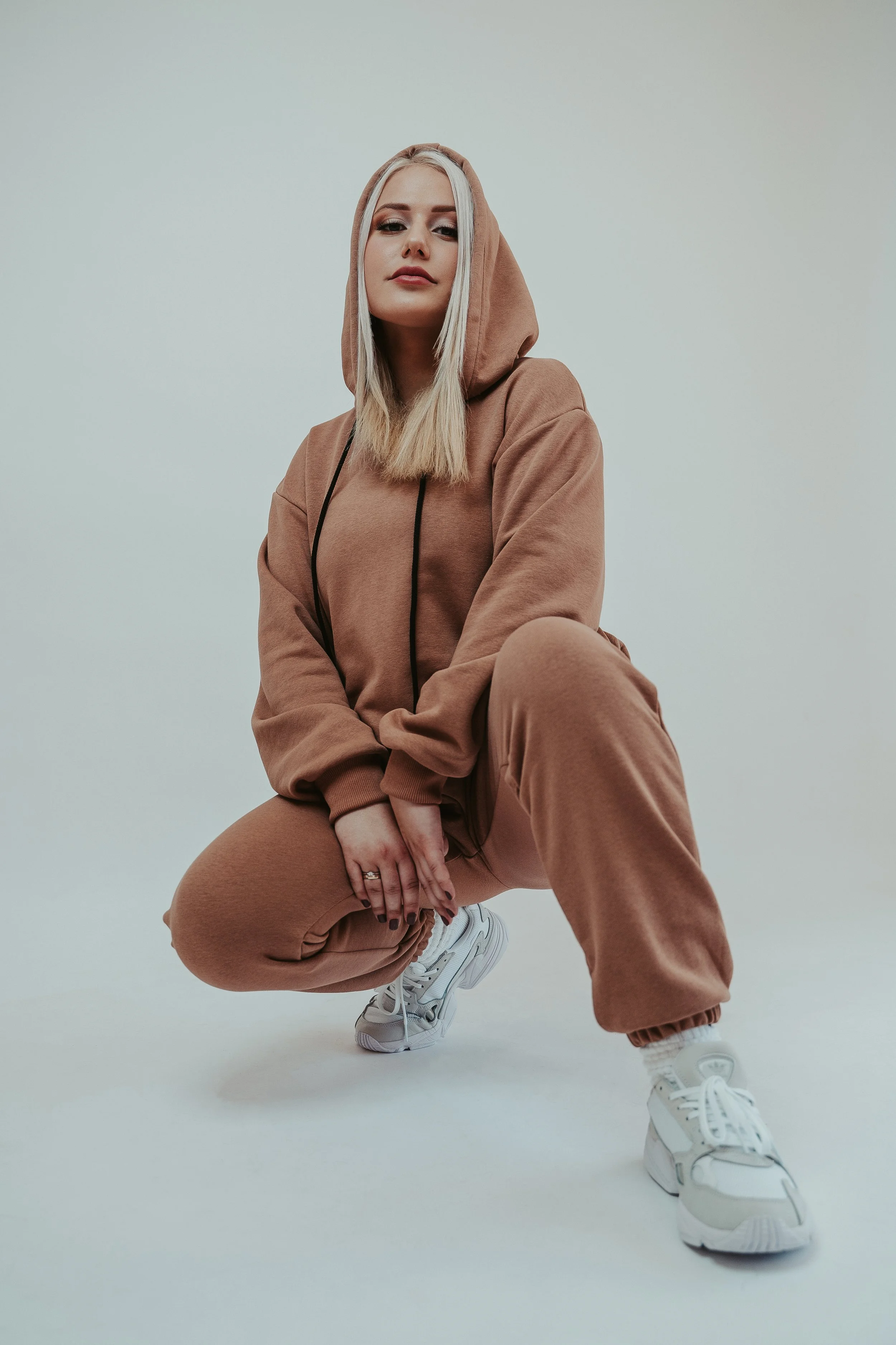 Young woman in tan hoodie and sweatpants, white athletic shoes, squatting with one knee bent and one foot flat, looking at camera with neutral expression against plain light background.