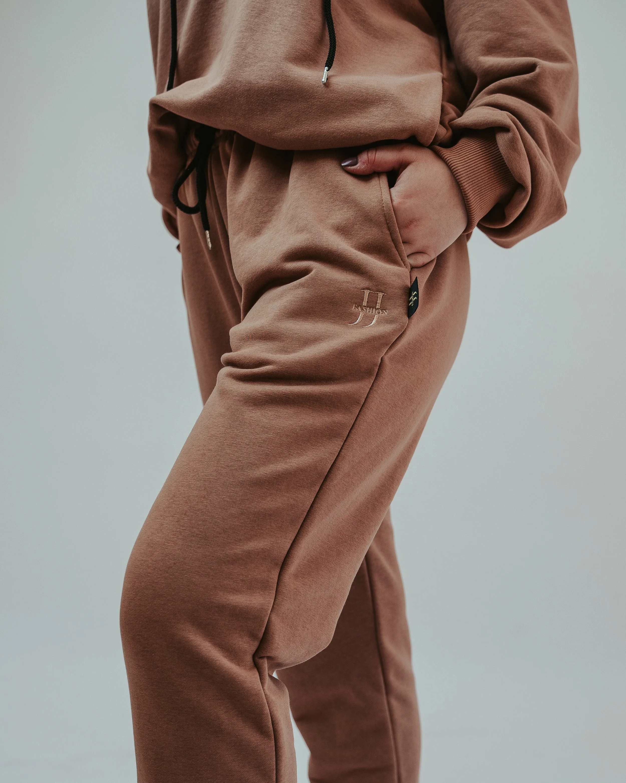 Close-up of a person wearing a brown hoodie and matching brown sweatpants with their right hand in the pocket.