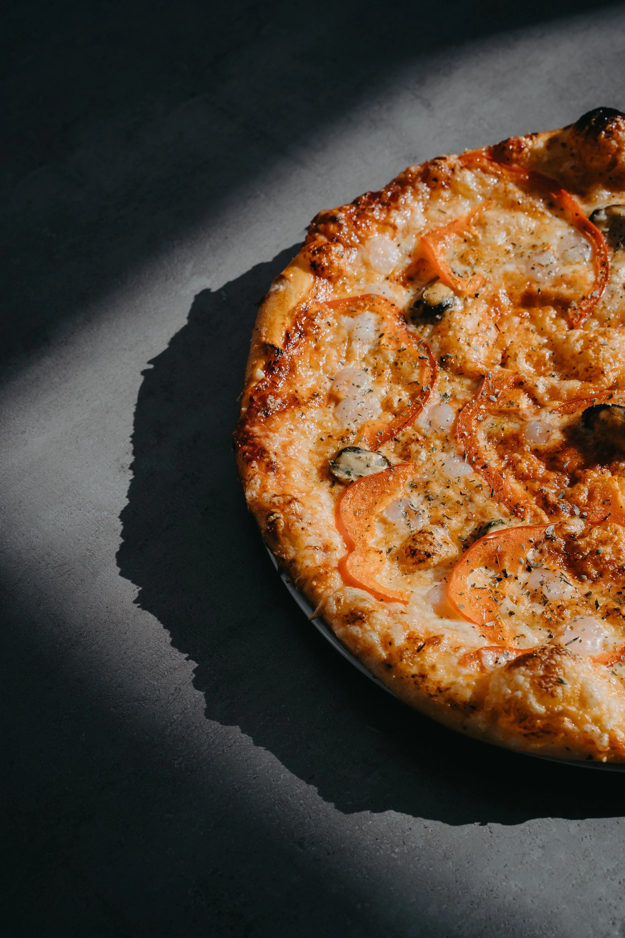Close-up of a cheese pizza with toppings, resting on a dark surface with shadow.