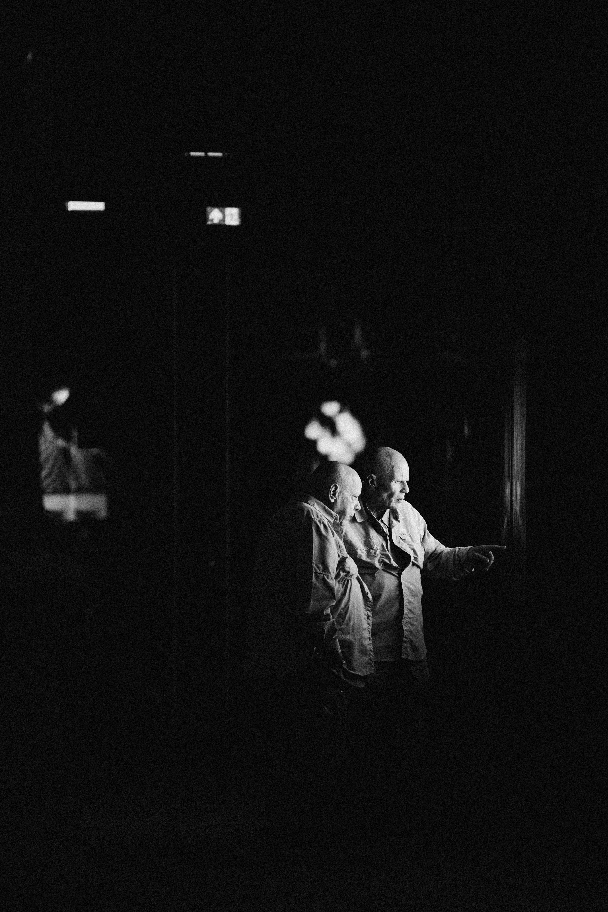 Two older men with bald heads are standing close together in a dark setting, with one of them pointing at something.