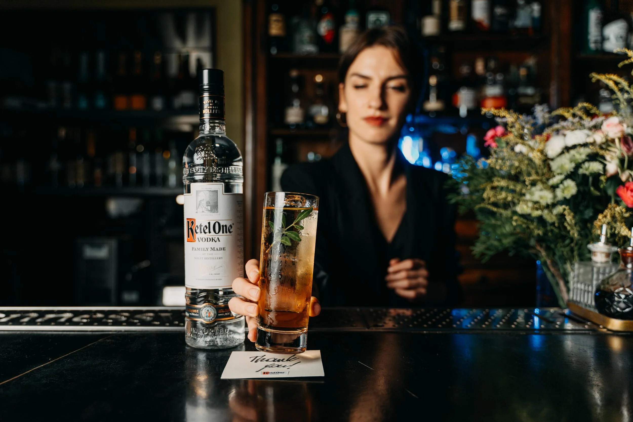 Bar scene with a woman in black holding a glass of drink garnished with herbs, a bottle of Ketel One vodka on the bar, and a bouquet of flowers nearby.