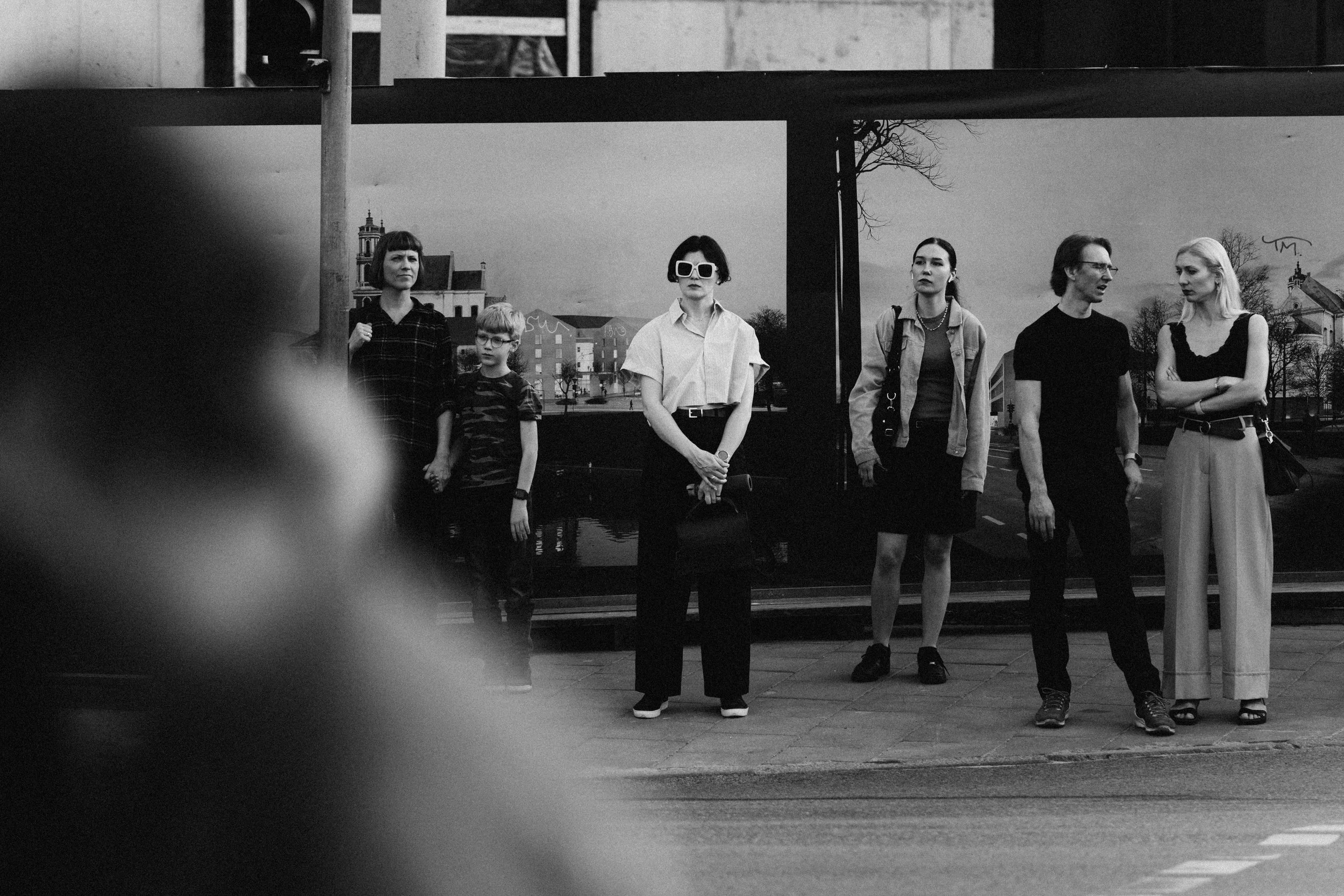 Black and white photo of six people standing on a sidewalk near a bus stop, with buildings and a church in the background.