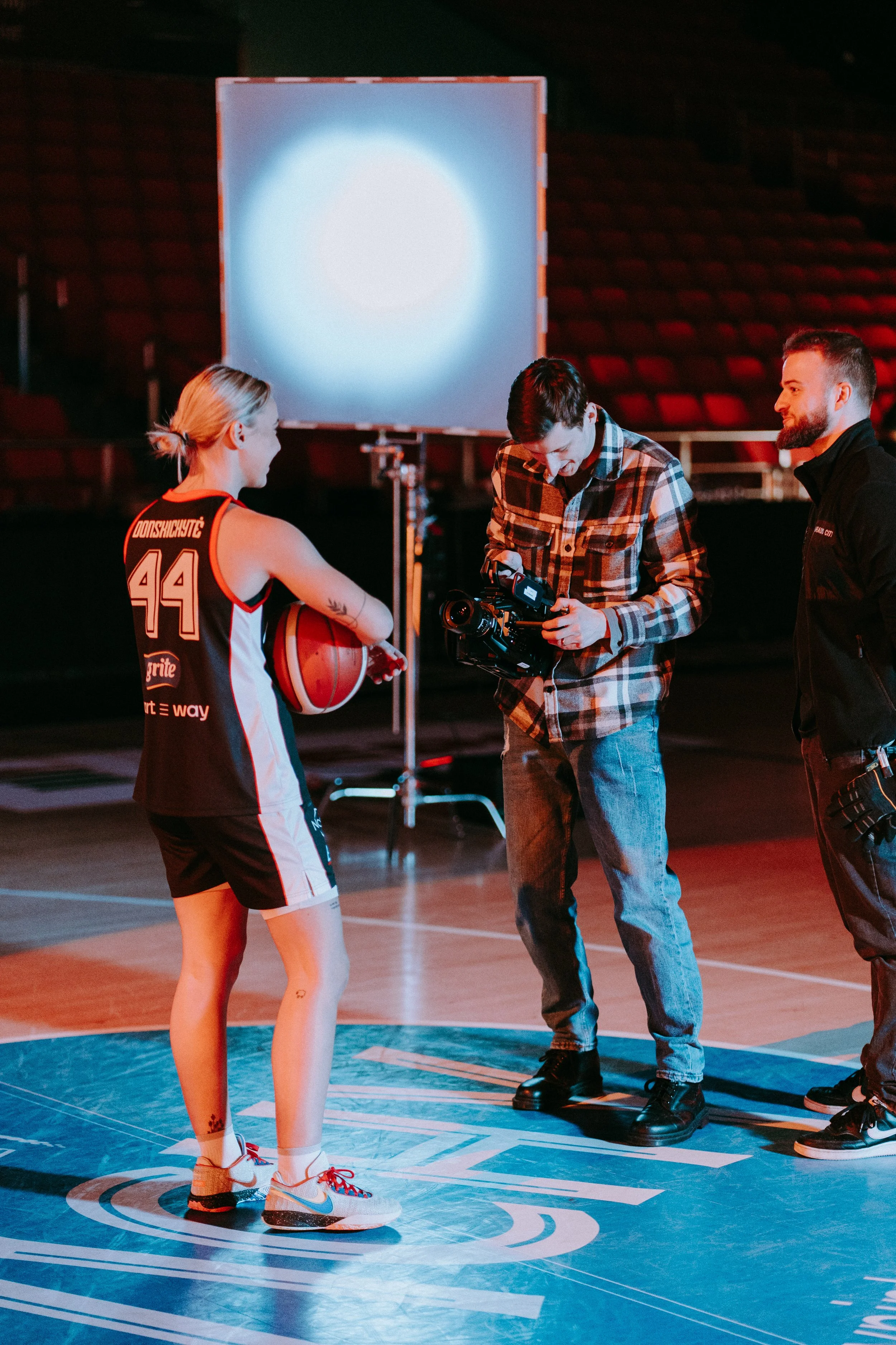 A female basketball player holding a basketball stands on a basketball court being filmed by two men, one operating a camera, during a professional shoot or interview.