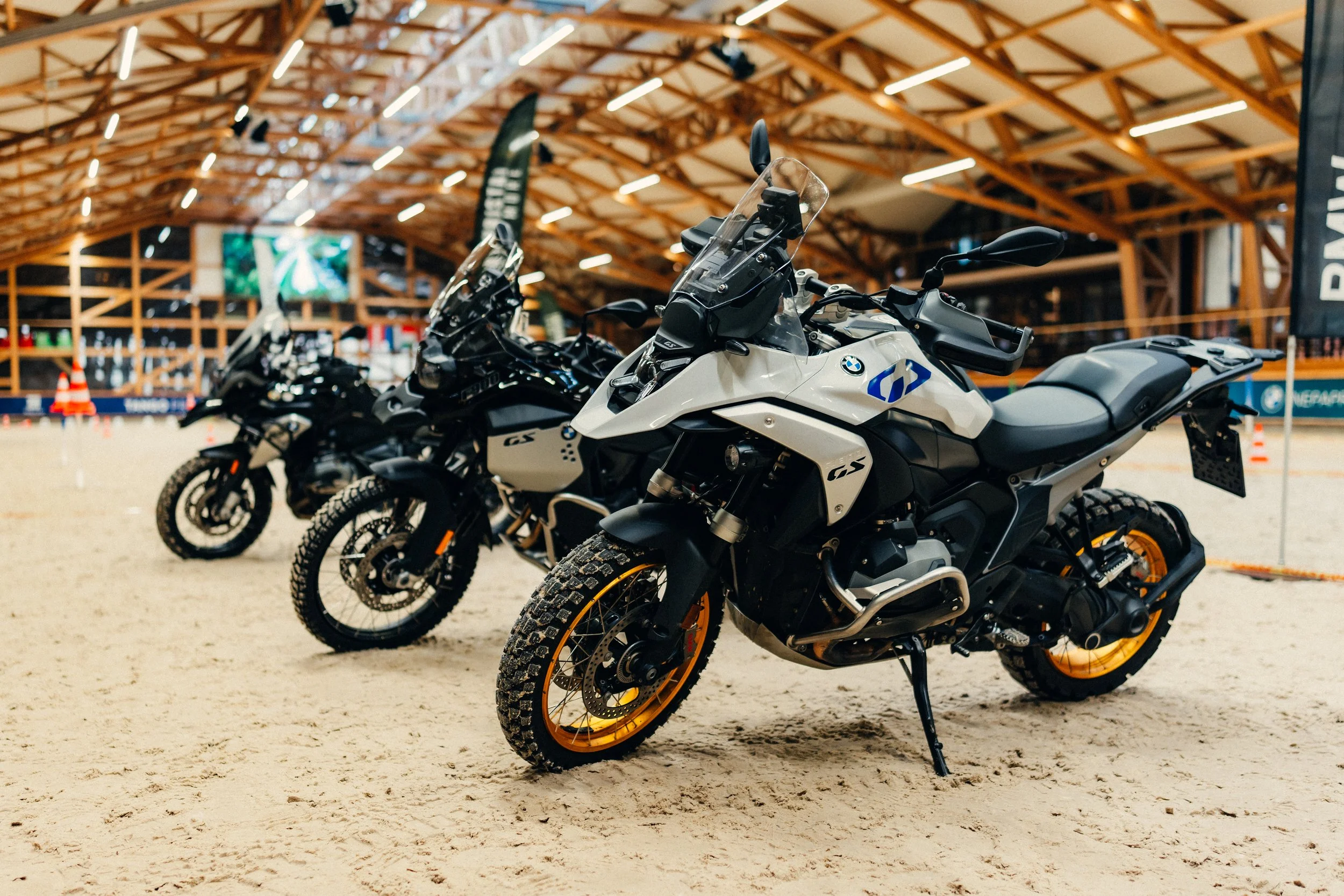 Three BMW adventure motorcycles lined up inside a large indoor space with a sandy floor, wooden ceiling beams, and banners.