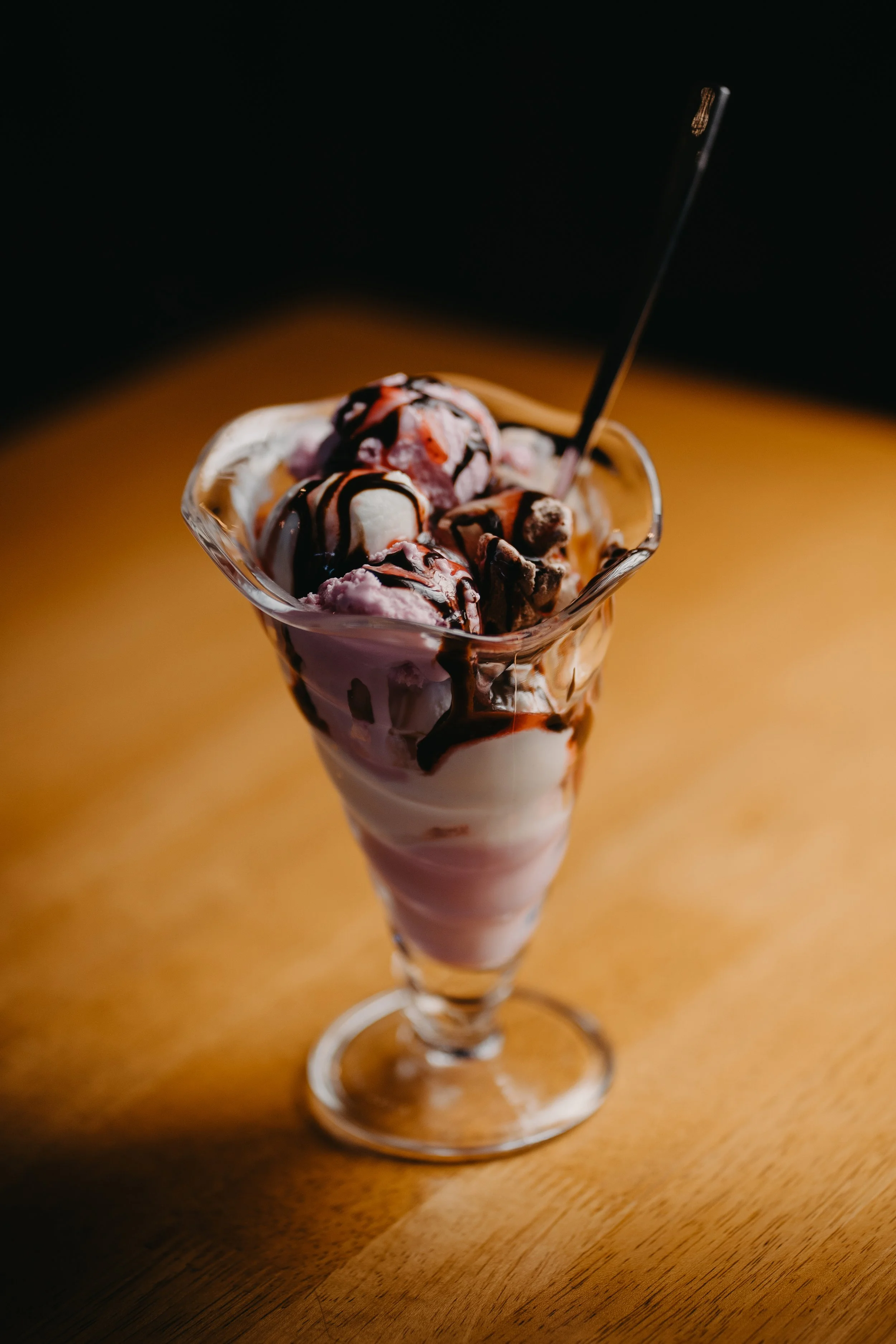 A tall glass of ice cream sundae topped with chocolate sauce, whipped cream, and various ice cream flavors on a wooden table.