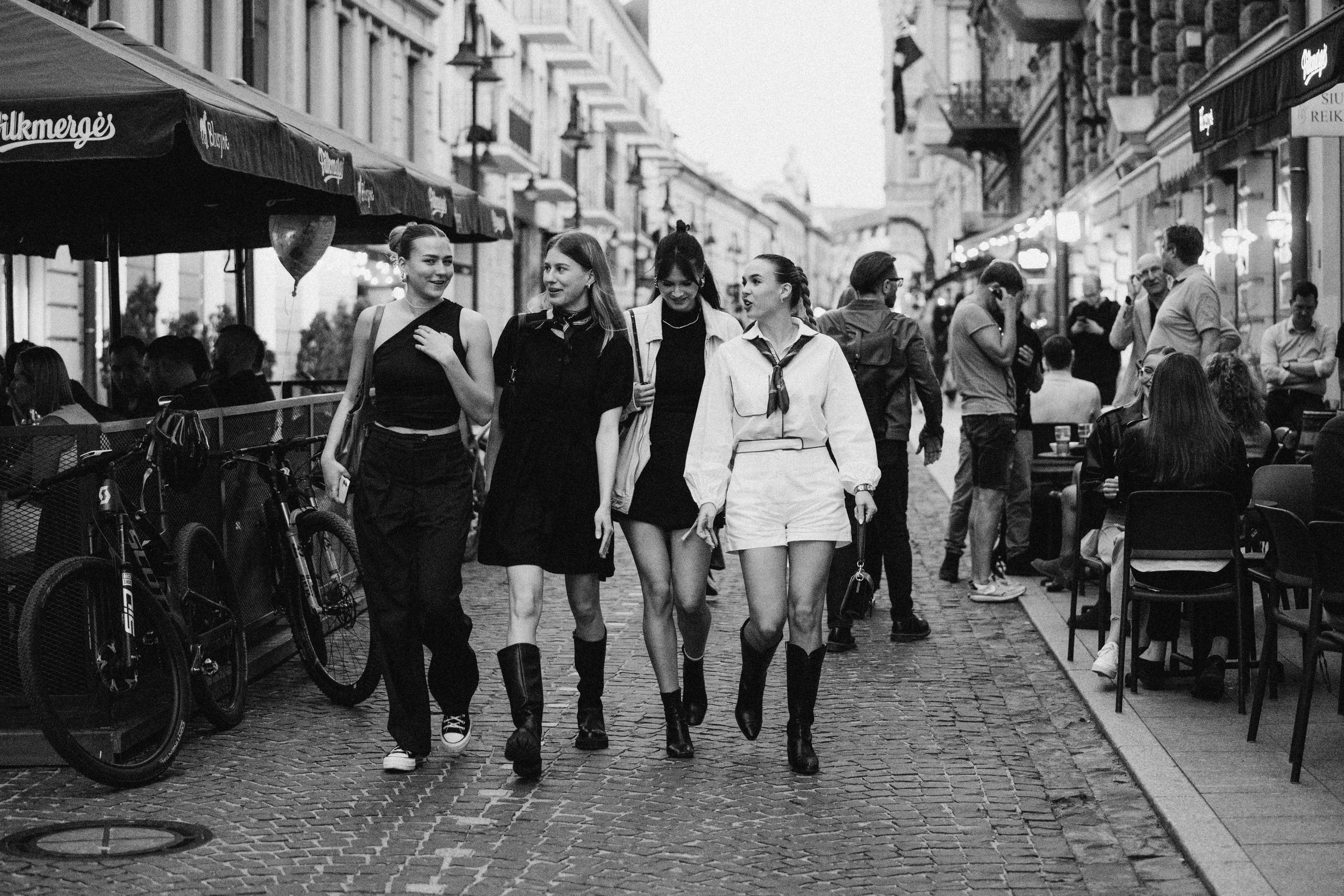 Four young women walking together on a cobblestone street in a city, with shops and cafes on either side, in black and white.