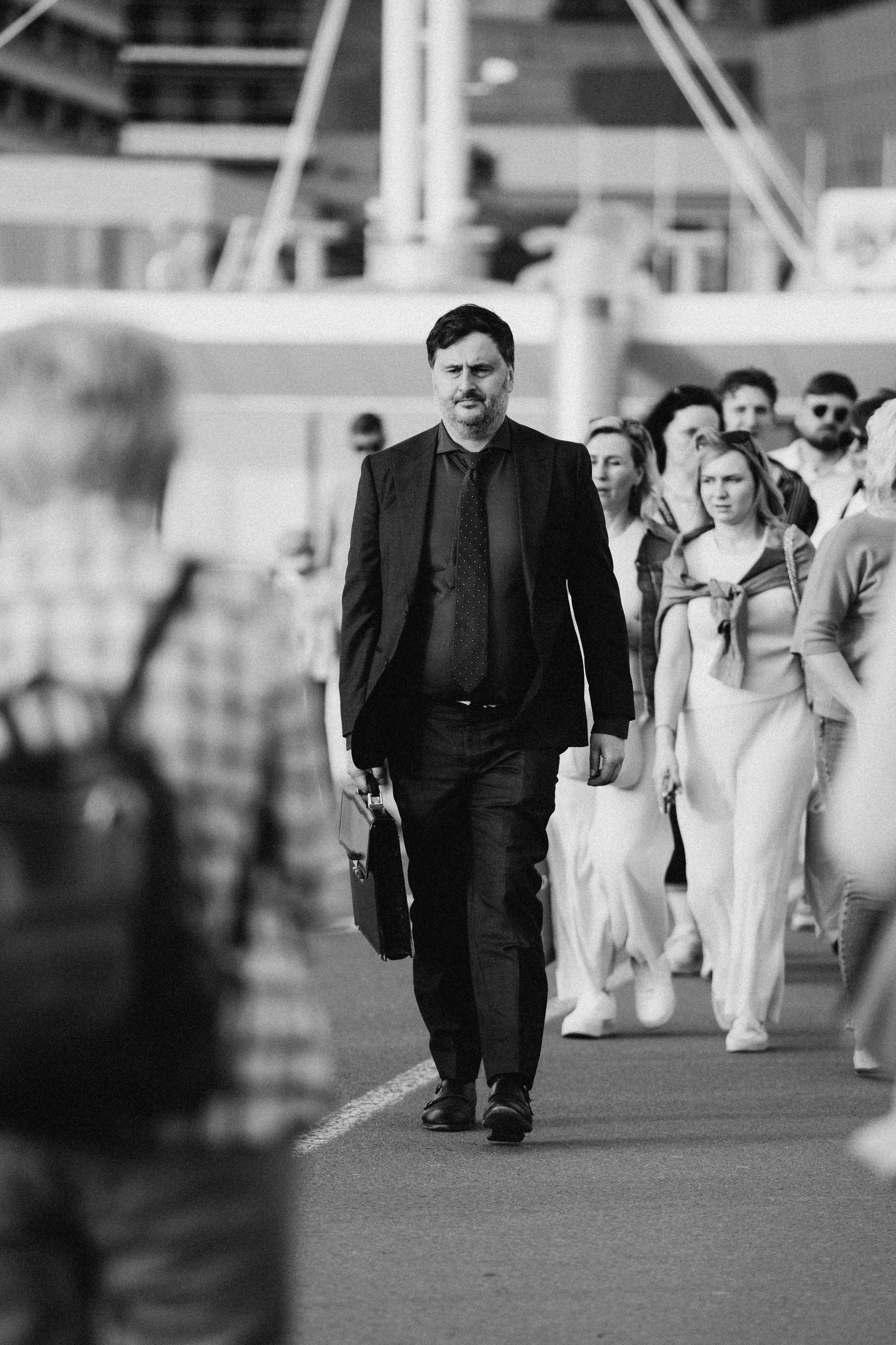 A man in a suit walking with a briefcase in front of a group of people at an airport or train station.