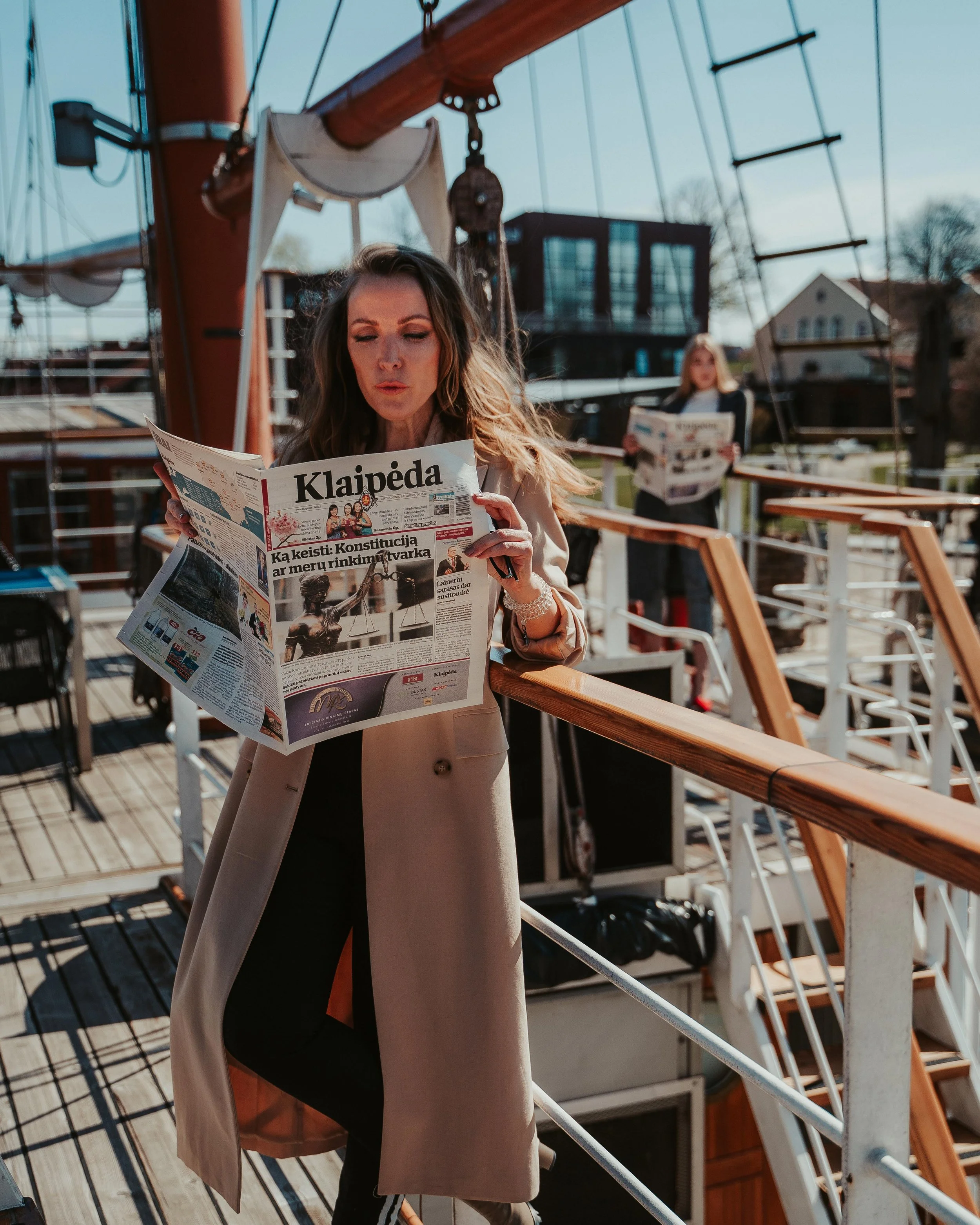 Two women reading newspapers on a boat deck with wooden railings and rigging, one in the foreground and the other in the background, during daytime.
