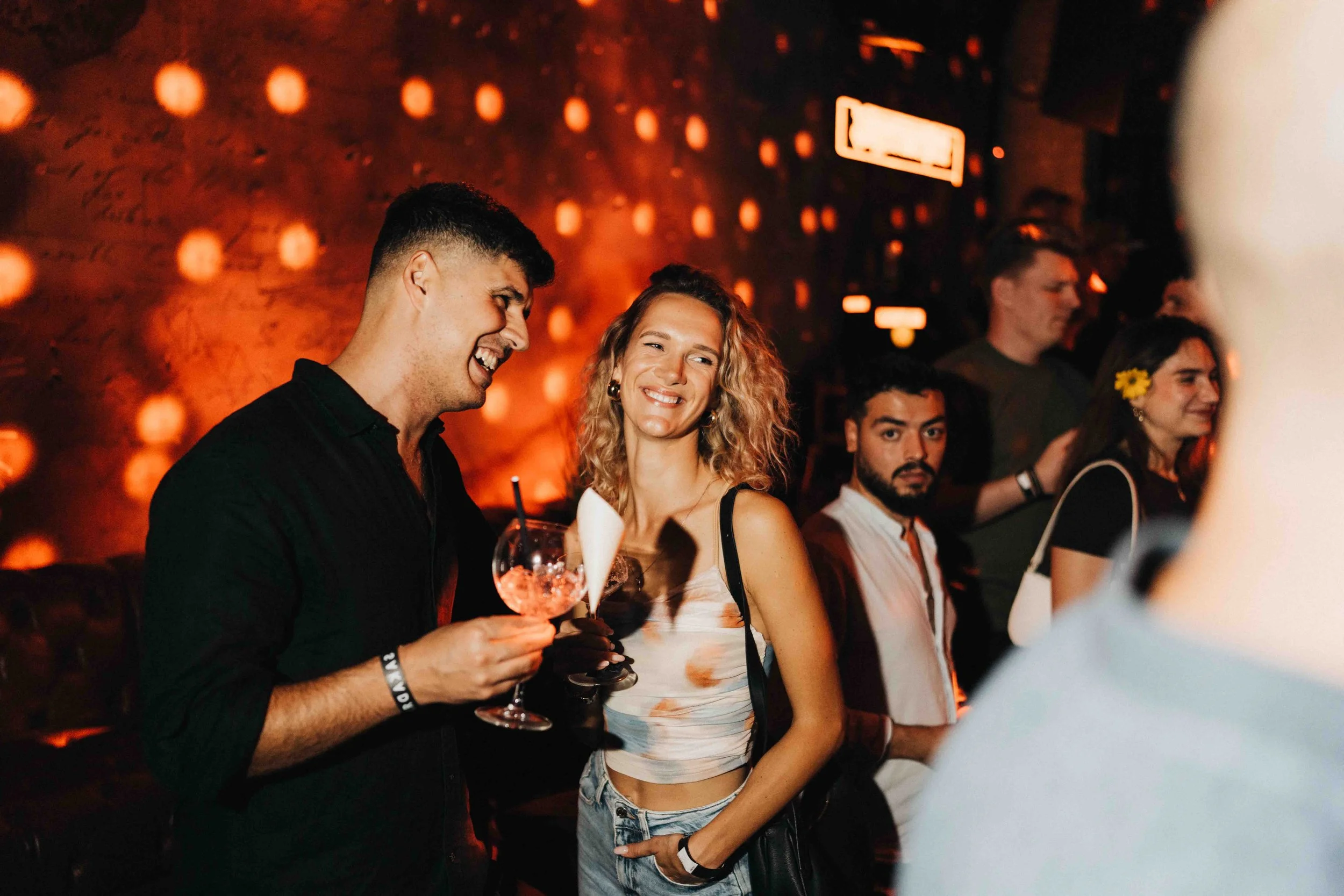 Group of people at a party or bar, smiling and holding cocktails, with warm, dim lighting and decorative background.