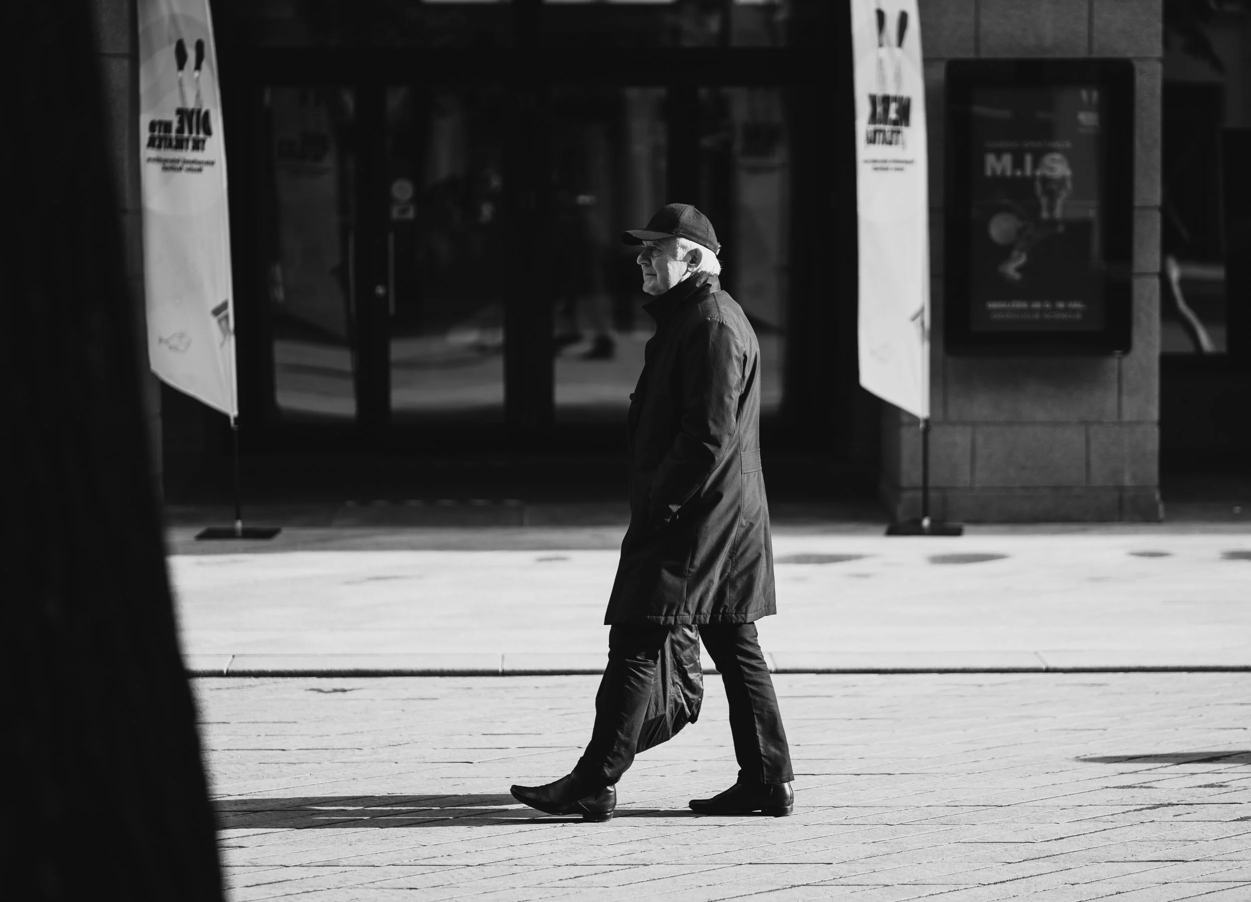 A man walking on a city sidewalk, dressed in a long coat and hat, with a building and signs in the background.