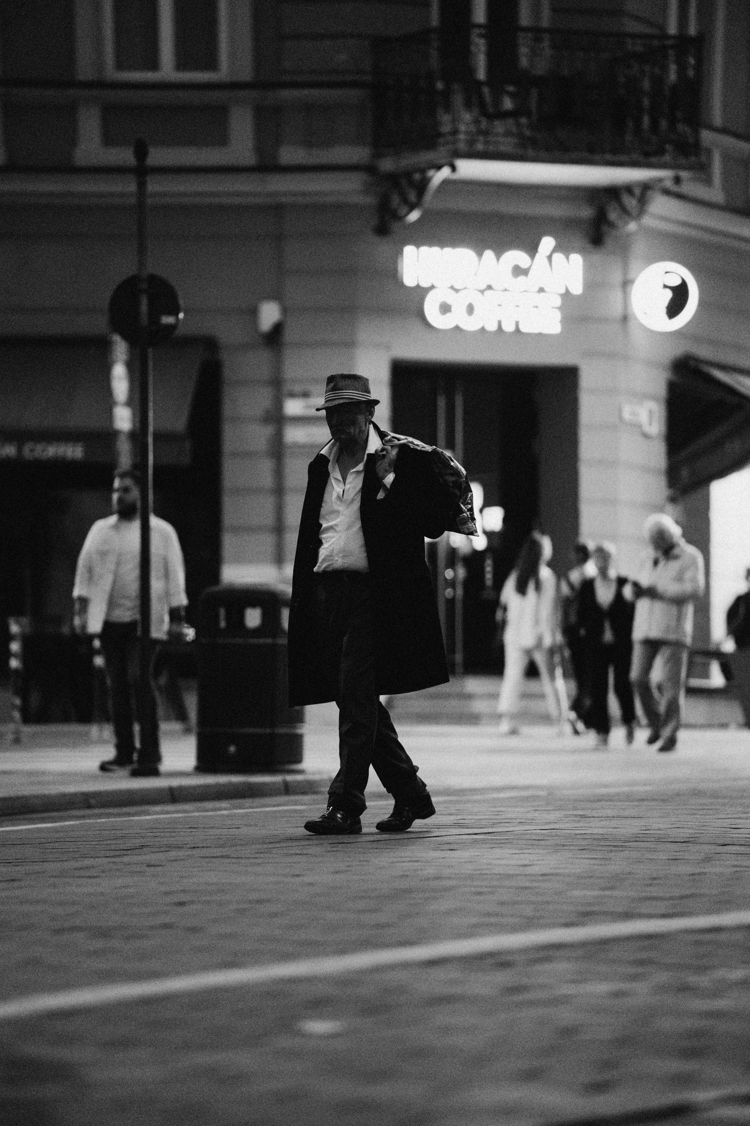 A black and white photo of a man in a coat and hat walking across an intersection in front of a coffee shop sign at night. Several pedestrians are visible in the background.