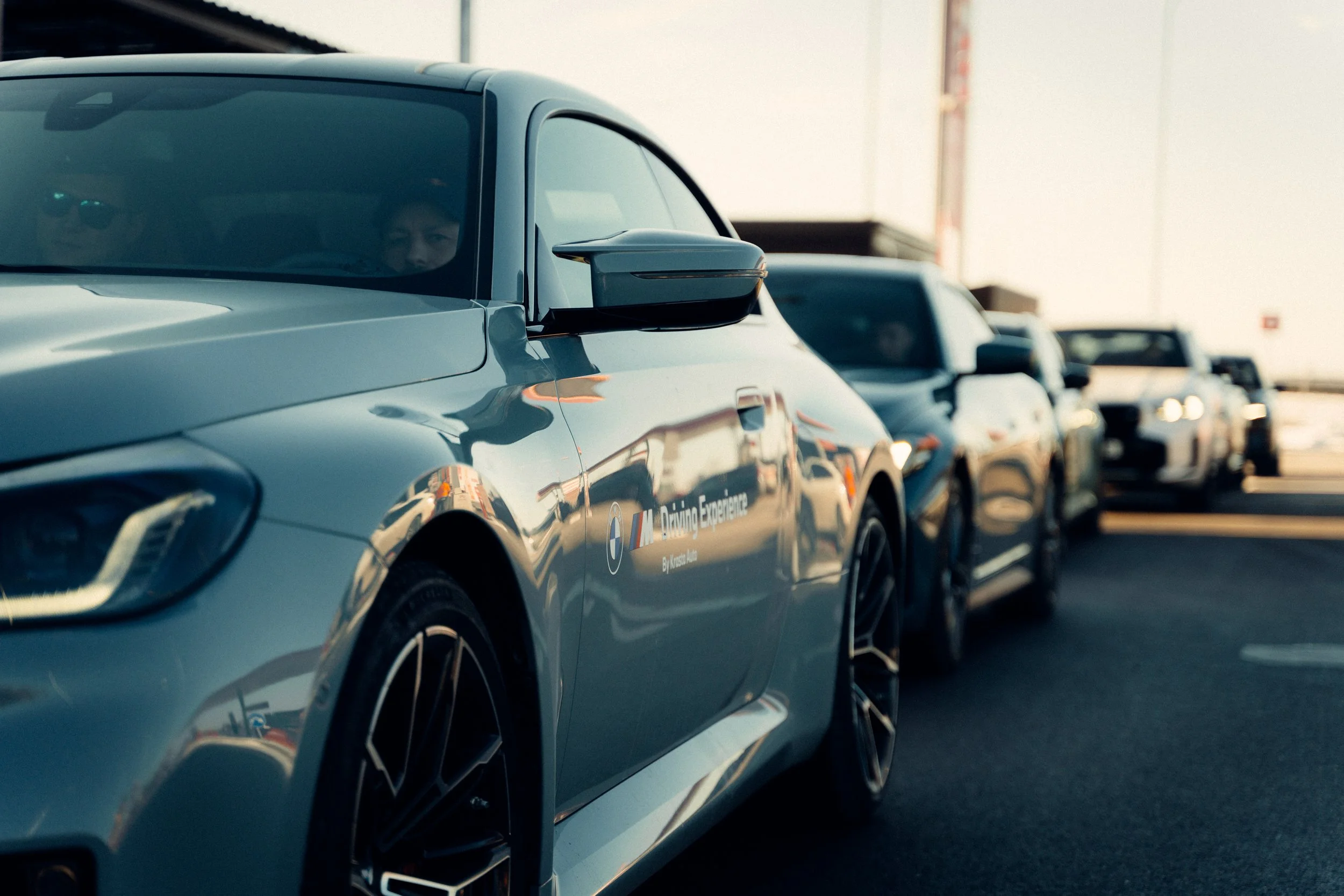 A line of luxury BMW cars parked on a road, with one car in the foreground displaying a 'Driving Experience' logo on the side, under a clear sky during sunset.