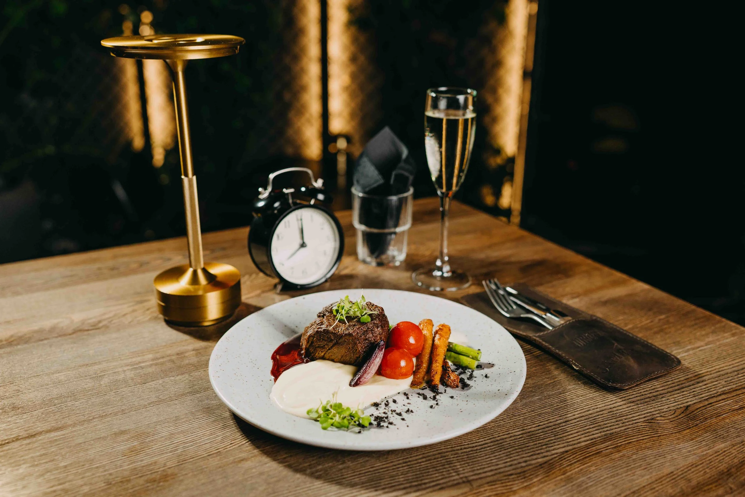 A prepared gourmet meal featuring steak, cherry tomatoes, green vegetables, and sauces on a white plate. In the background are a glass of white wine, an alarm clock, a glass with a black napkin, silverware on a leather napkin holder, and a gold decor