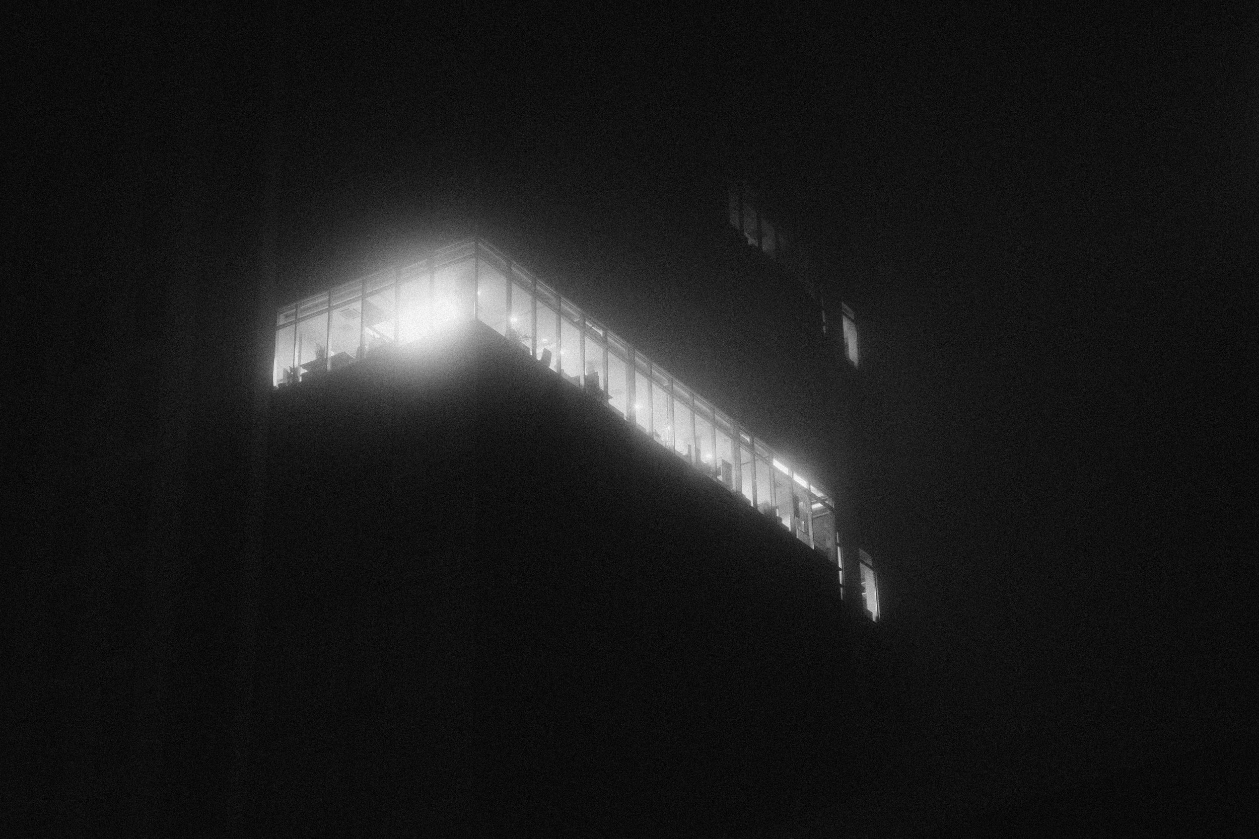 A black and white photo of a modern building at night with illuminated windows, creating a stark contrast against the dark sky.