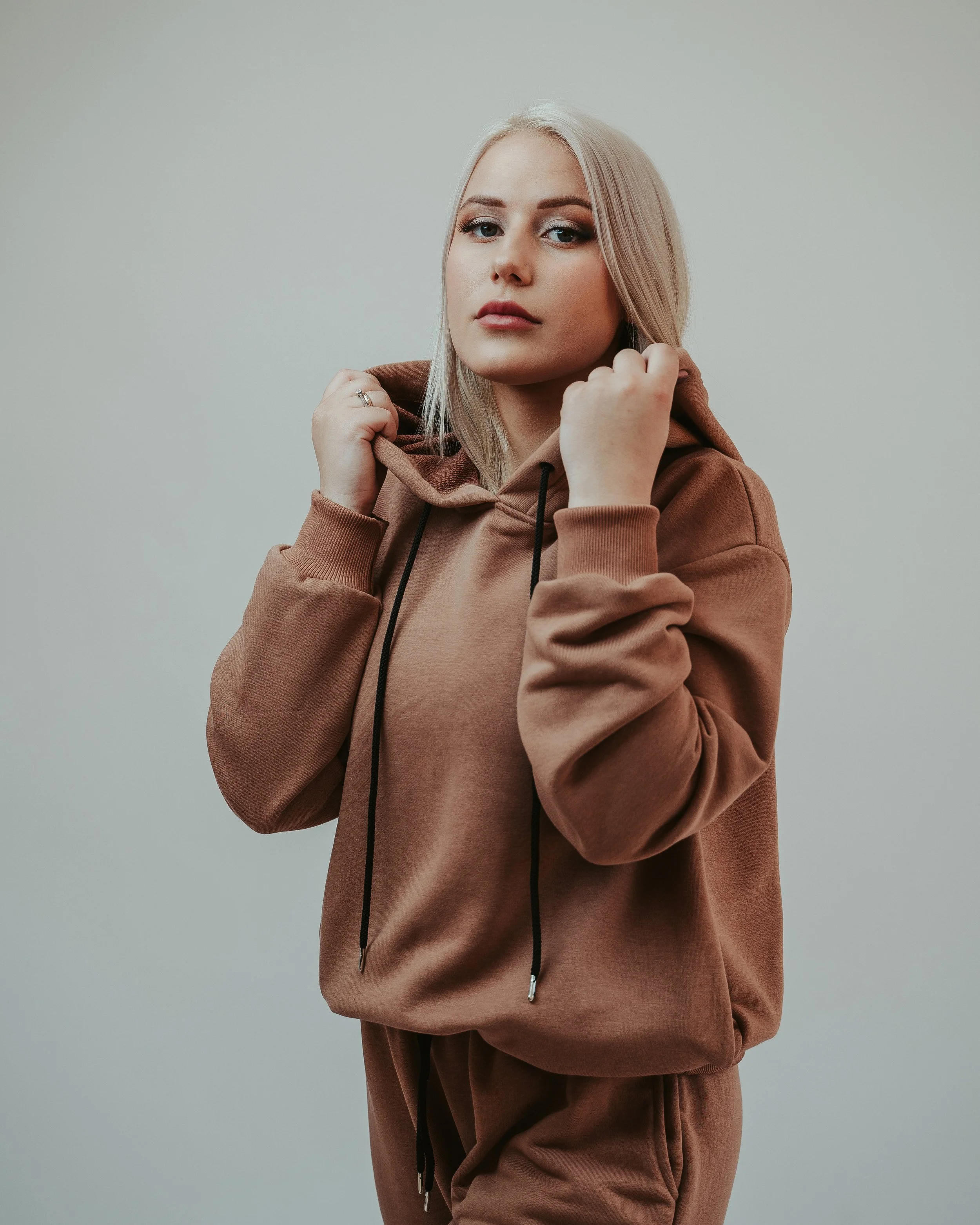 A young woman with blonde hair wearing a brown hoodie and matching sweatpants, holding the hoodie’s drawstrings, standing against a plain, light-colored background.