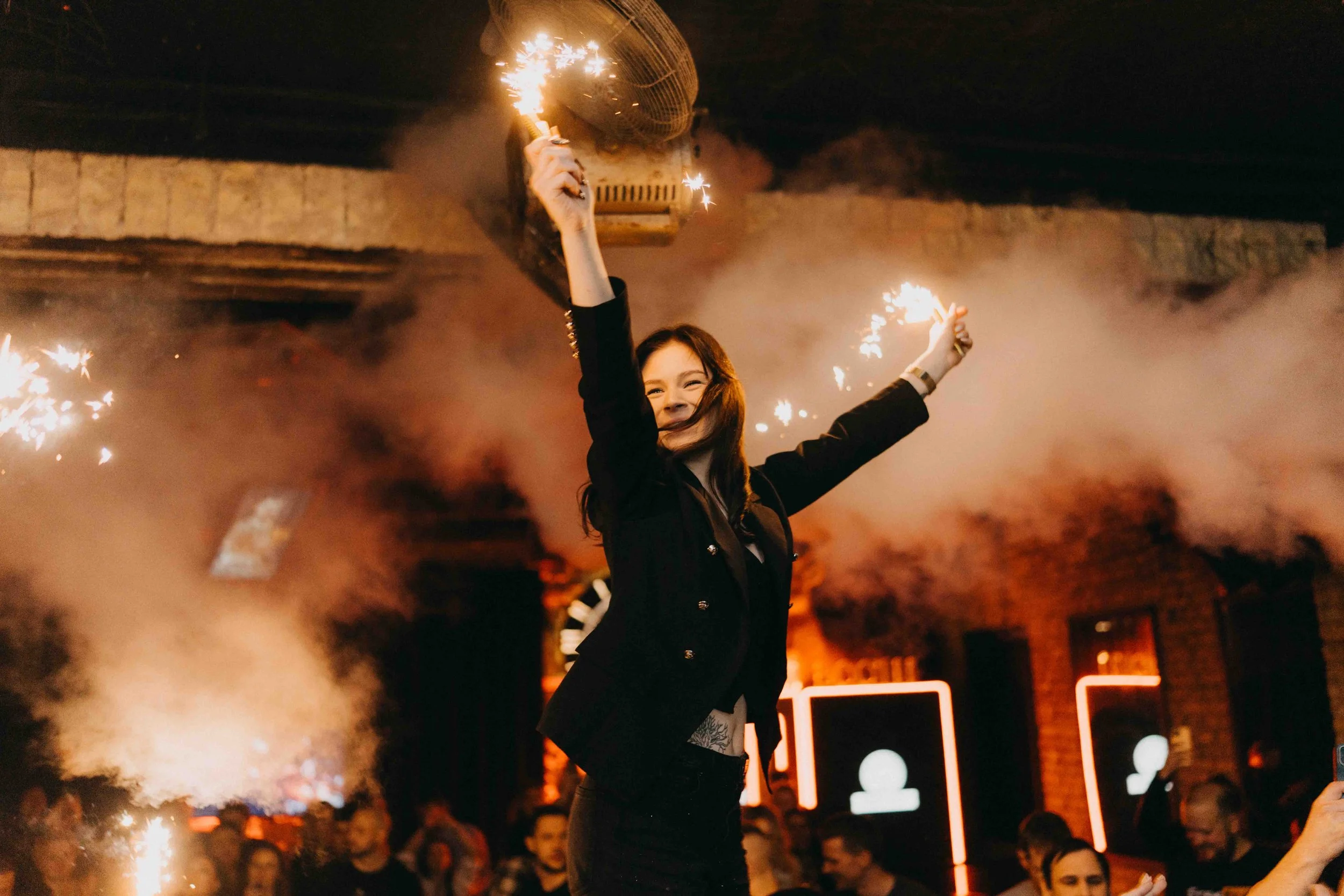 A woman celebrating with sparklers in a lively indoor party atmosphere, with smoke effects and many people in the background.