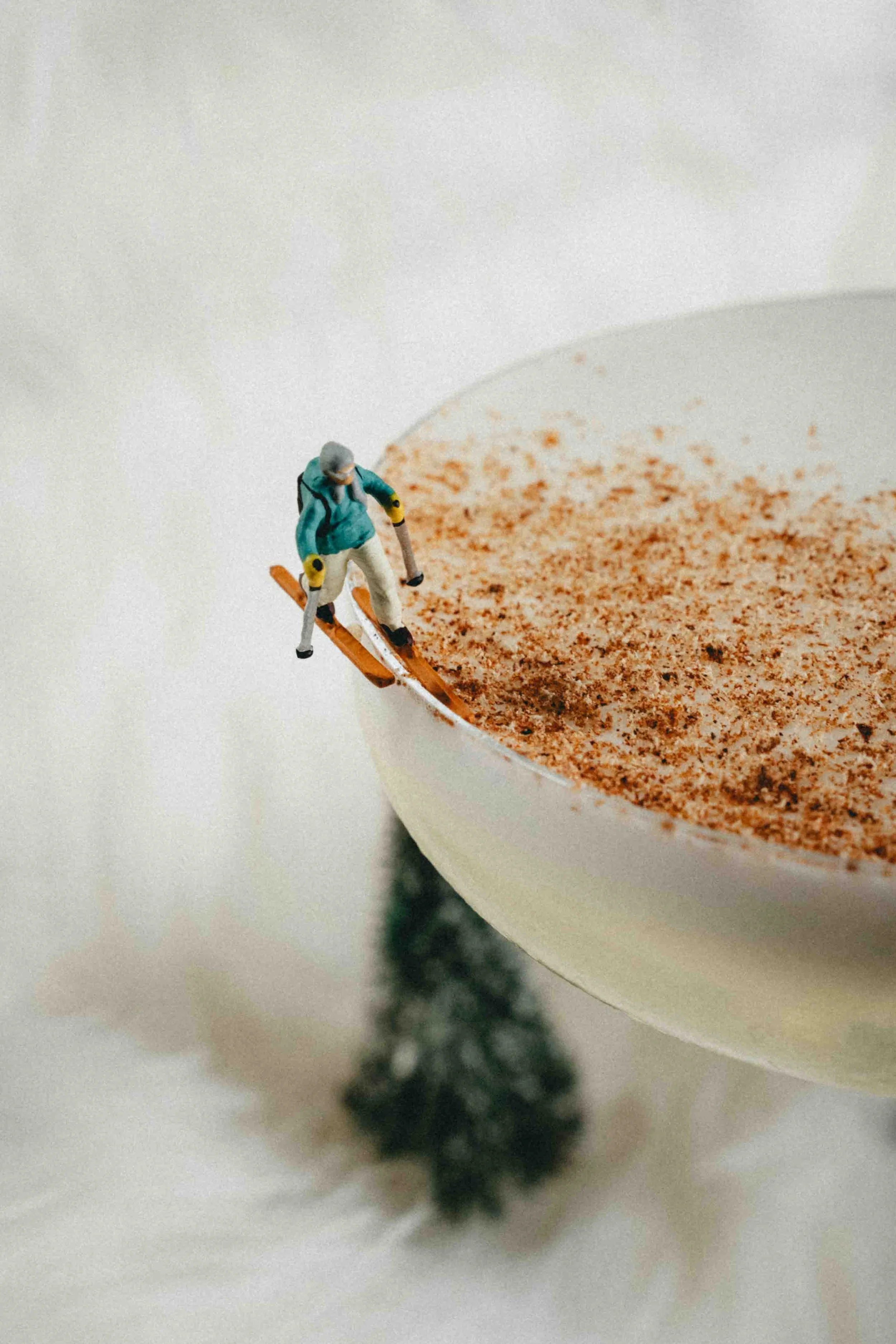 Miniature figurine of a skier skiing on the edge of a cup filled with a cinnamon or cocoa powder, with a blurred tree-like object below.