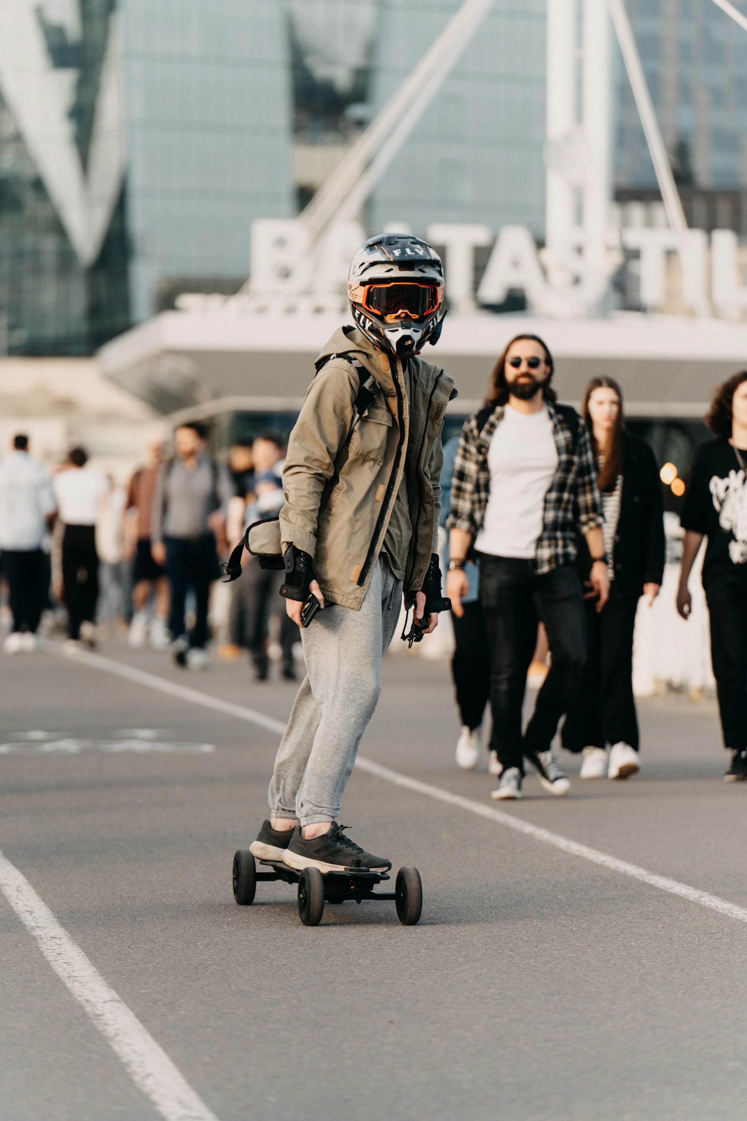 Person wearing a helmet and jacket riding a skateboard on a city street, with a group of people walking nearby and a building with a large sign in the background.