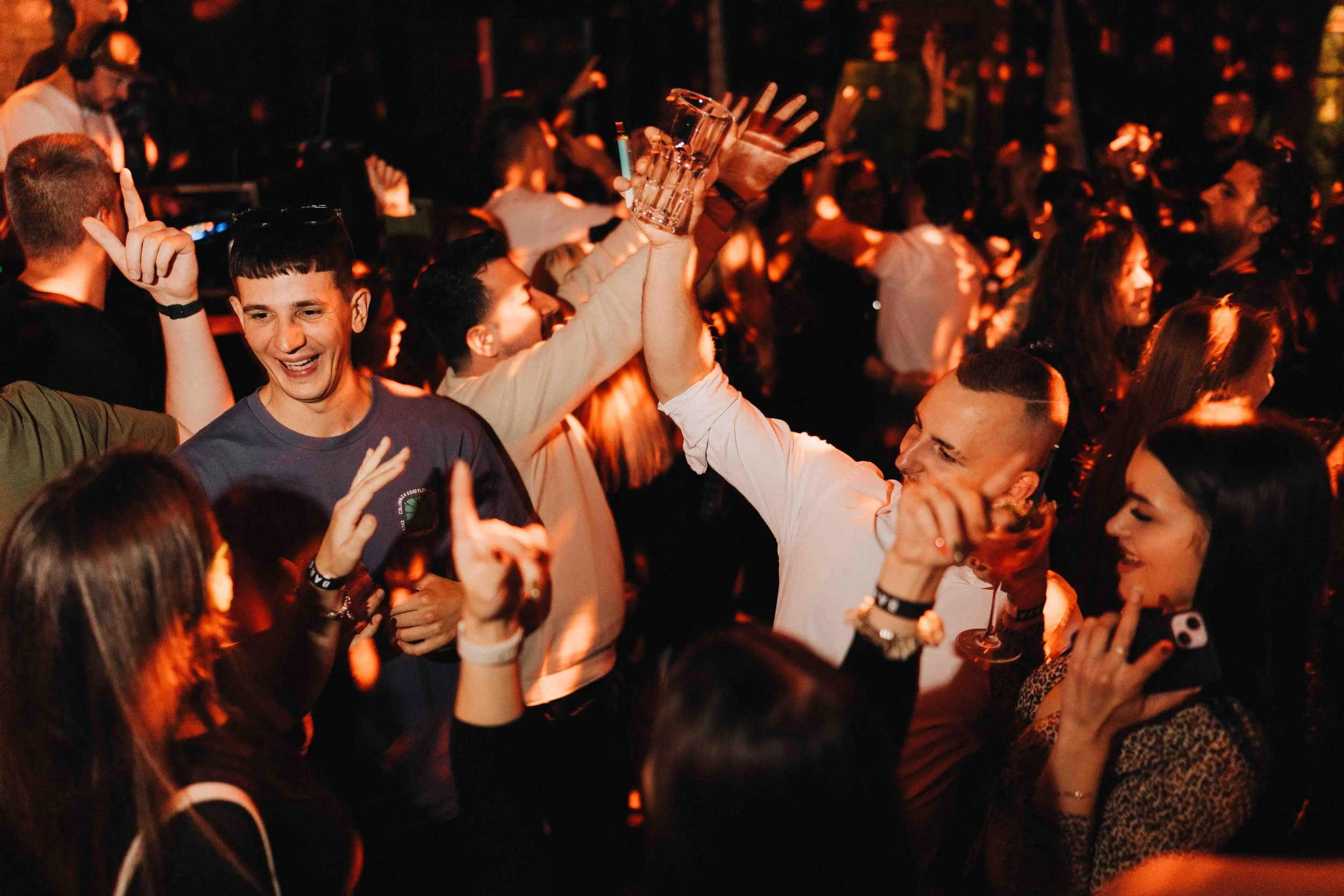 Group of people dancing and celebrating at a lively party or nightclub, with some raising their hands and holding drinks.