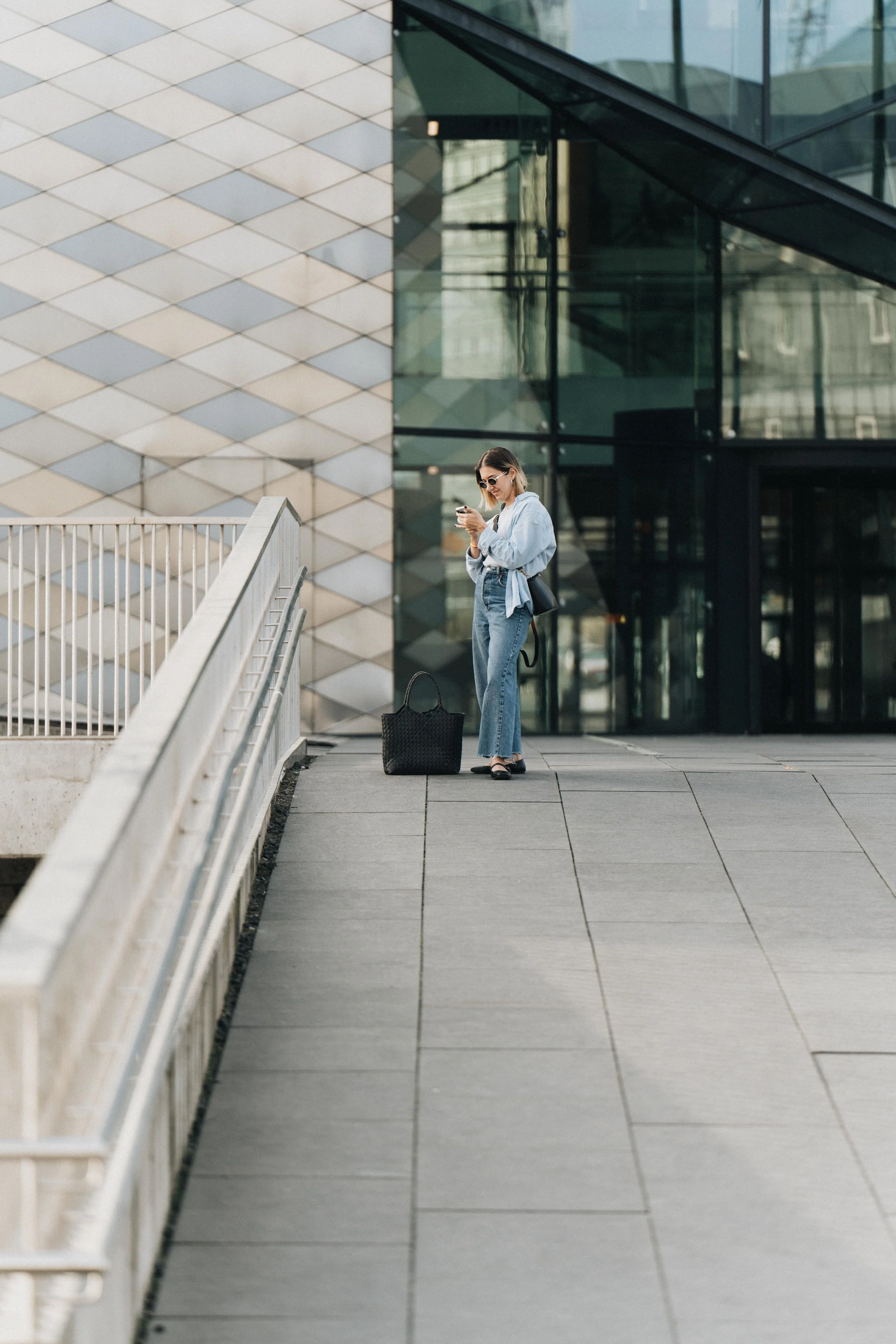 A woman with sunglasses and a denim jacket standing on an outdoor walkway in front of a modern glass building, looking at her phone with a black bag on the ground beside her.