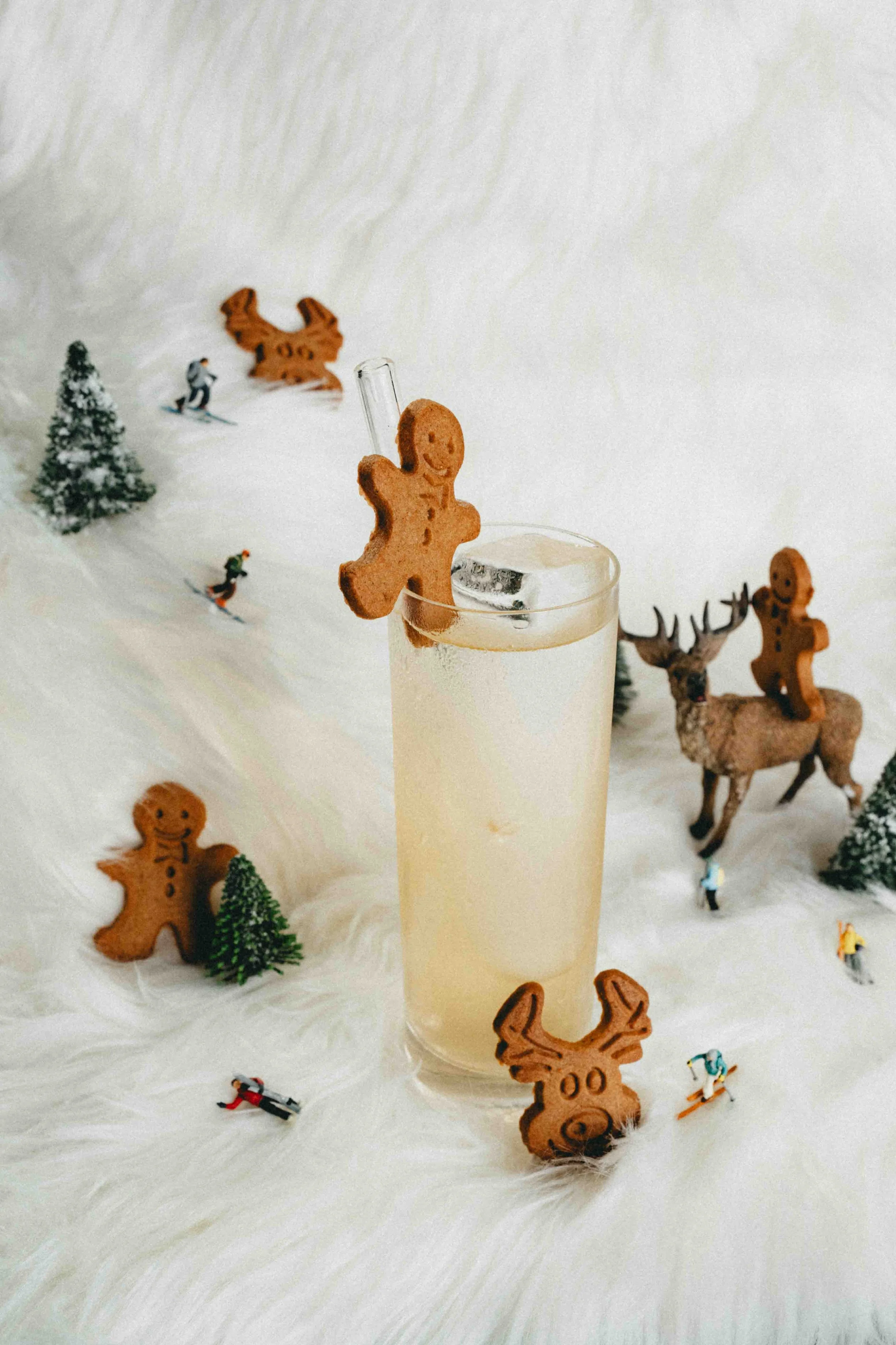 A beverage in a tall glass with a gingerbread man cookie hanging on the rim. The background includes miniature Christmas trees, reindeer, snow-covered figurines, and gingerbread cookies on a white furry surface.