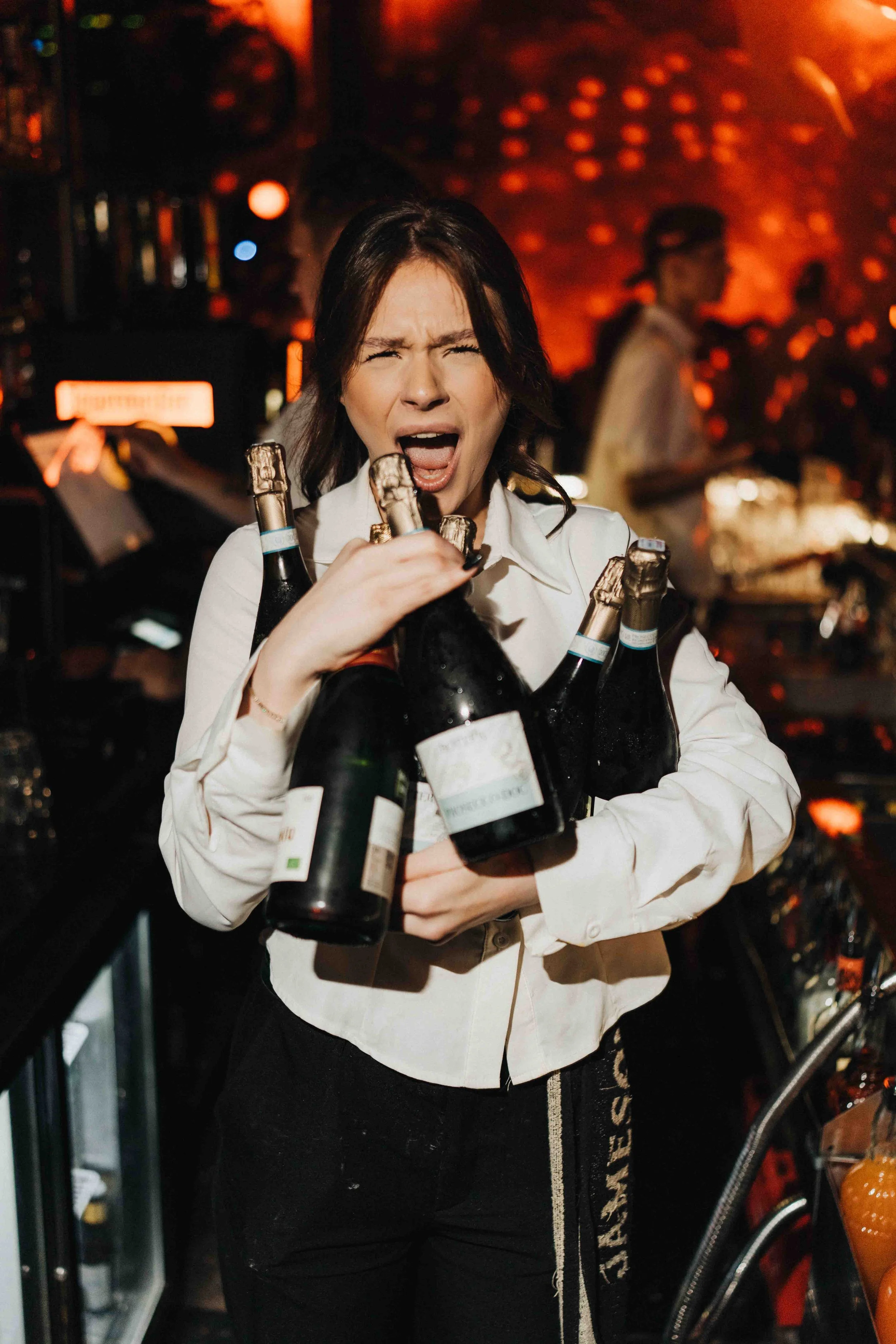 A woman with a distressed expression holding multiple bottles of champagne in a bar or nightclub setting with a fiery background.
