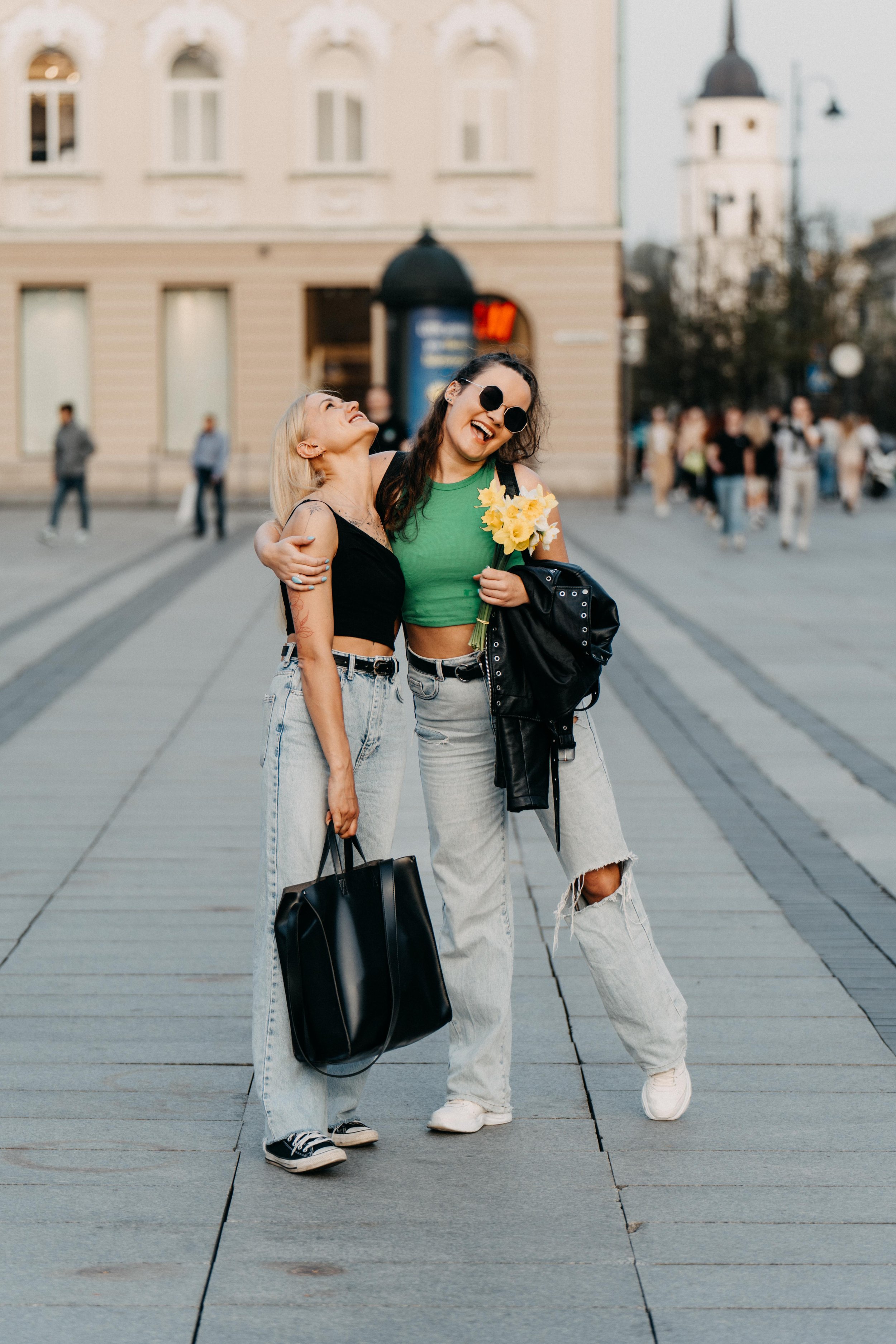 Two young women are standing together on a city street, smiling and laughing. One woman has blonde hair and is wearing a black crop top, light jeans, and black sneakers. The other woman has dark hair, is wearing sunglasses, a green crop top, ripped j