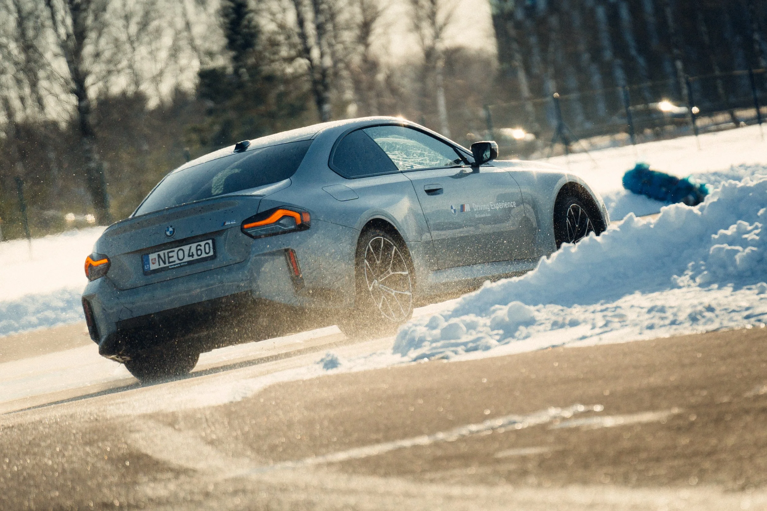 A silver BMW car driving on a snow-covered road during winter, kicking up snow behind it.